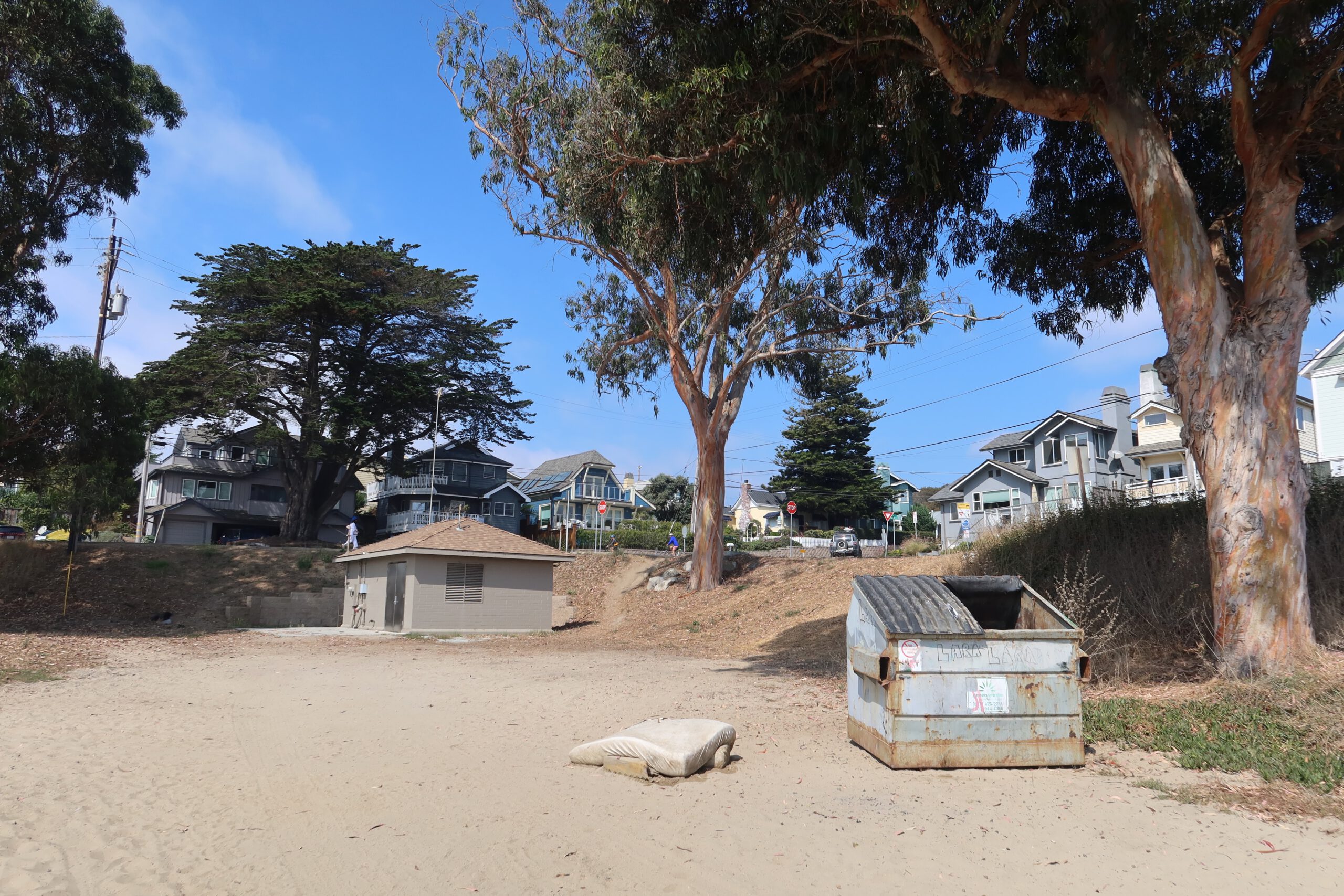 Beach access area near Twin Lakes State Beach in Santa Cruz with coastal homes, eucalyptus trees, a dumpster, and a discarded mattress on the sand