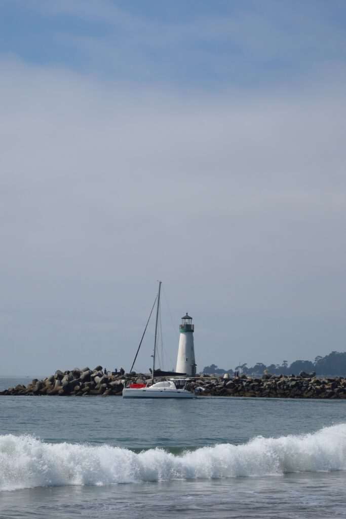 Santa Cruz Harbor lighthouse near Twin Lakes State Beach with a sailboat passing the rock jetty and waves breaking in the foreground