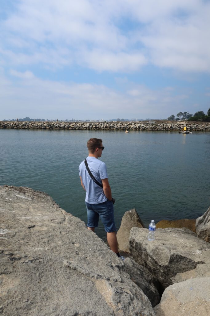 Person standing on coastal rocks near Twin Lakes State Beach in Santa Cruz, California overlooking calm harbor waters and the rock jetty