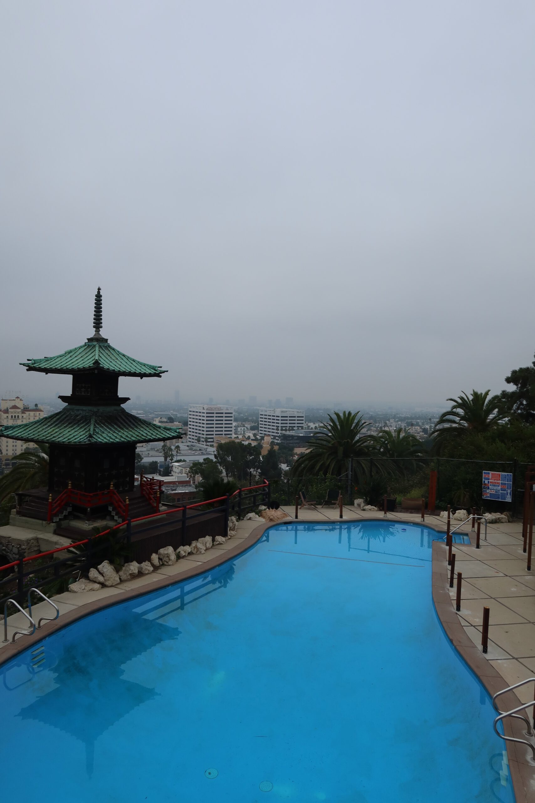 Infinity-style swimming pool at a Hollywood Hills hotel overlooking Los Angeles, with palm trees, a pagoda-style structure, and city skyline visible in the distance under an overcast sky.