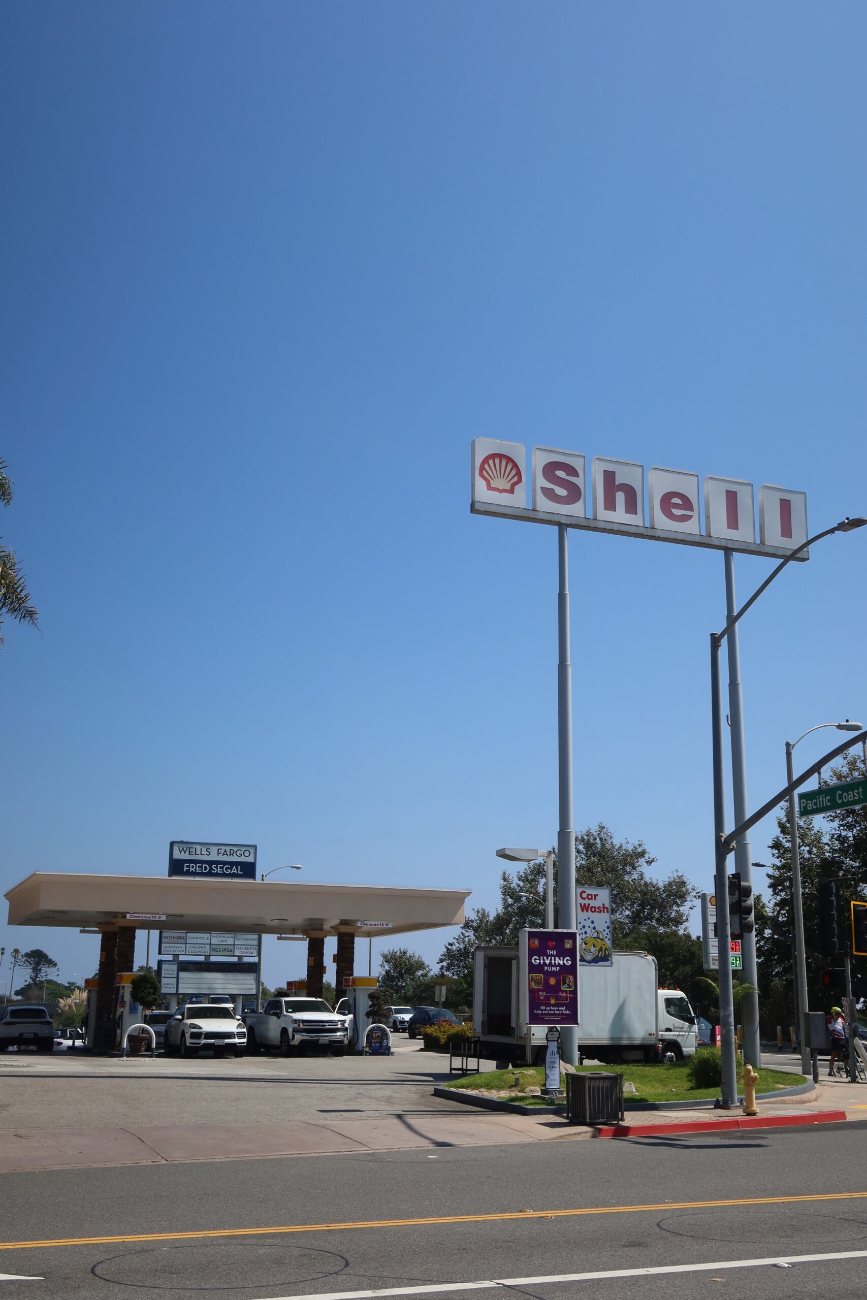 Shell gas station along Pacific Coast Highway in Malibu, California, with the classic Shell sign against a clear blue sky, fuel pumps in use, and Malibu Country Mart visible in the background on a sunny day.