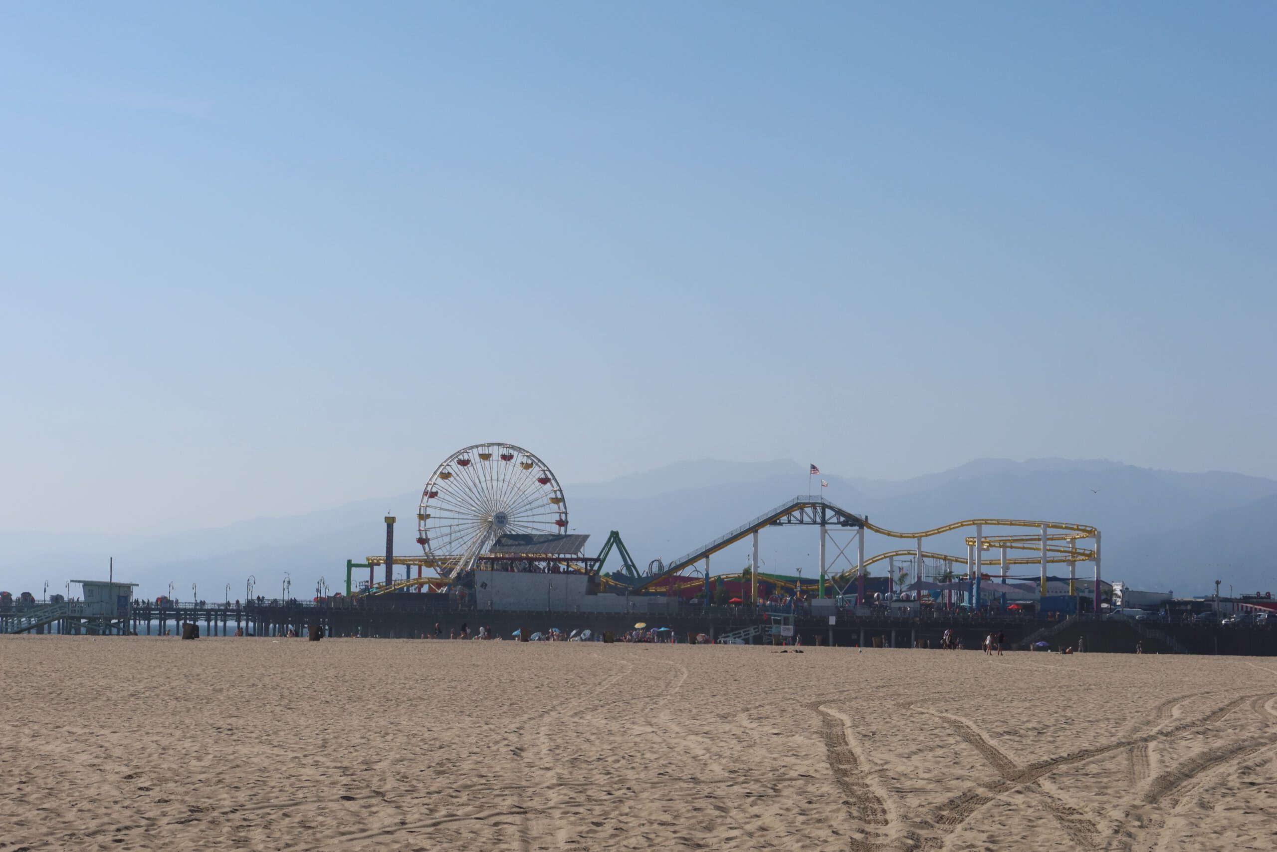 Wide beach view of Santa Monica Pier with Pacific Park’s Ferris wheel and roller coaster rising above the pier, soft mountain silhouettes in the background, clear blue sky overhead, and sandy foreground with visible tire tracks and beachgoers in the distance.