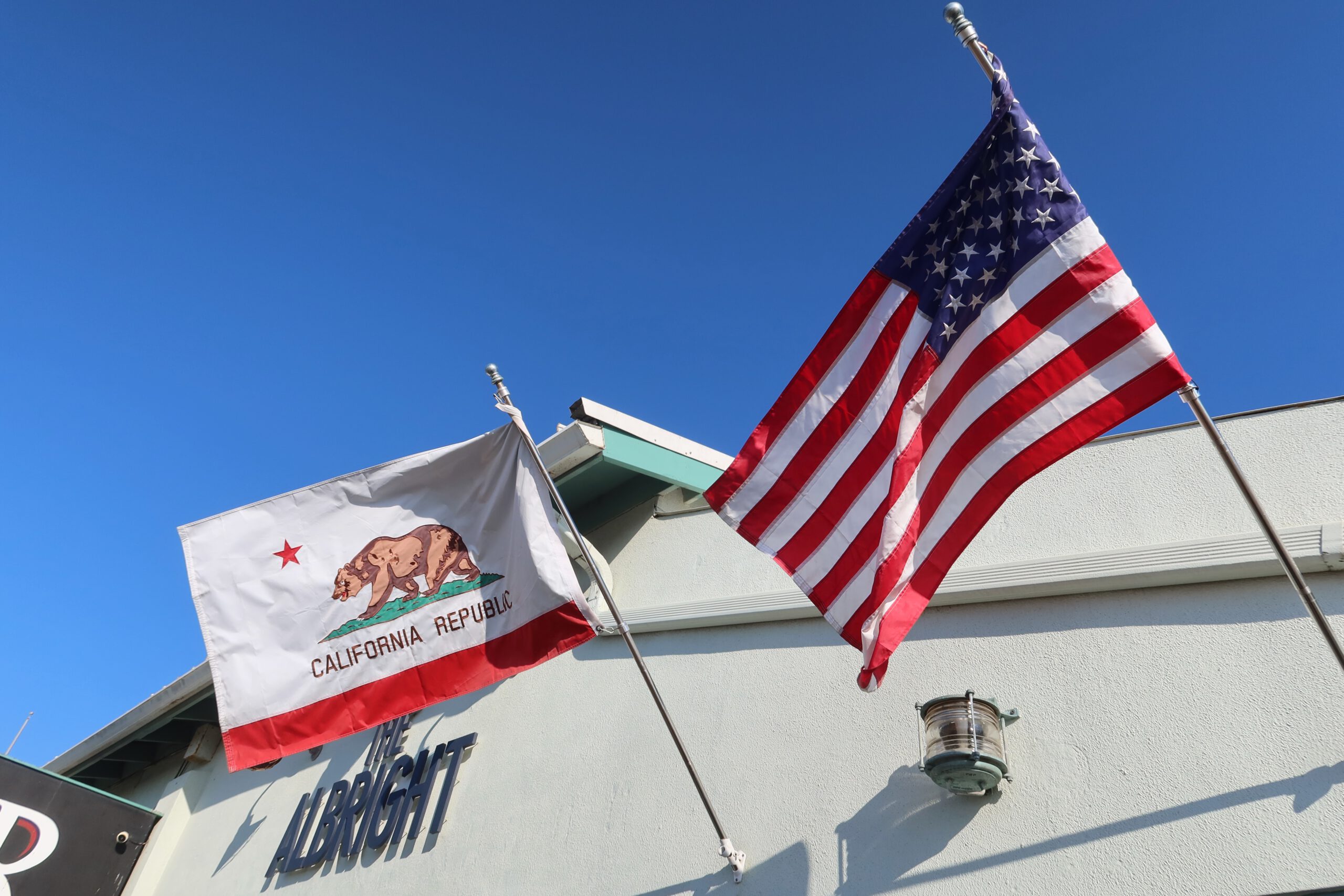 American flag and California Republic state flag waving side by side above pier buildings at Santa Monica Pier, captured on a sunny day with a deep blue California sky.