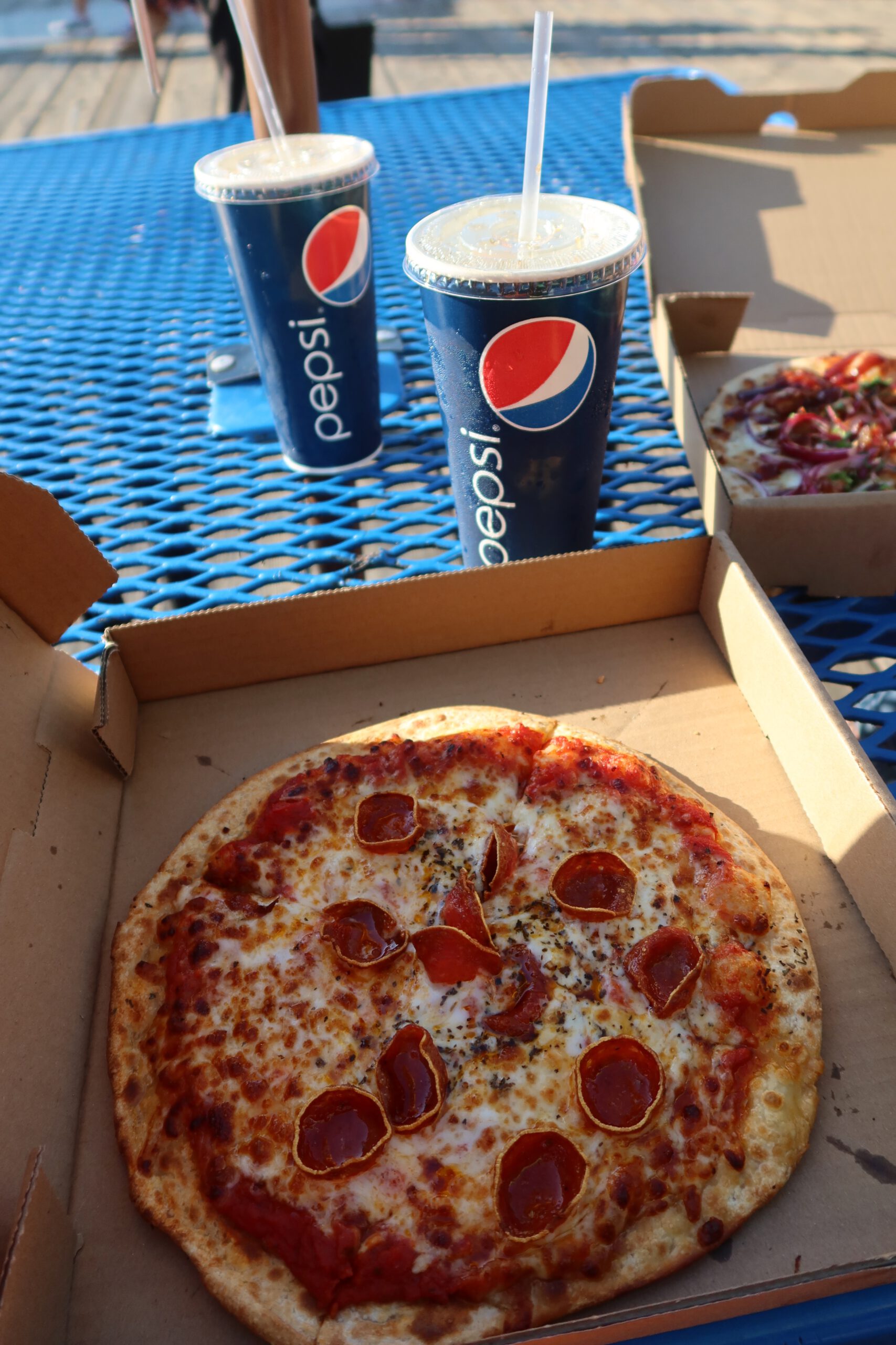 Fresh pepperoni pizza from Mr. Nice Fry at Santa Monica Pier served in a cardboard box, with large American-sized Pepsi sodas on a blue outdoor picnic table near the beach.