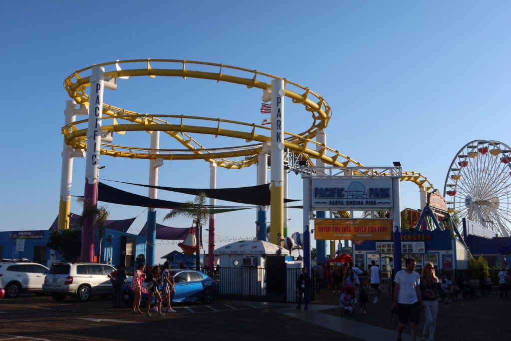 Entrance view of Pacific Park on Santa Monica Pier with the yellow West Coaster roller coaster looping overhead, colorful park signage, visitors walking across the pier, and the iconic Ferris wheel visible against a clear blue California sky.