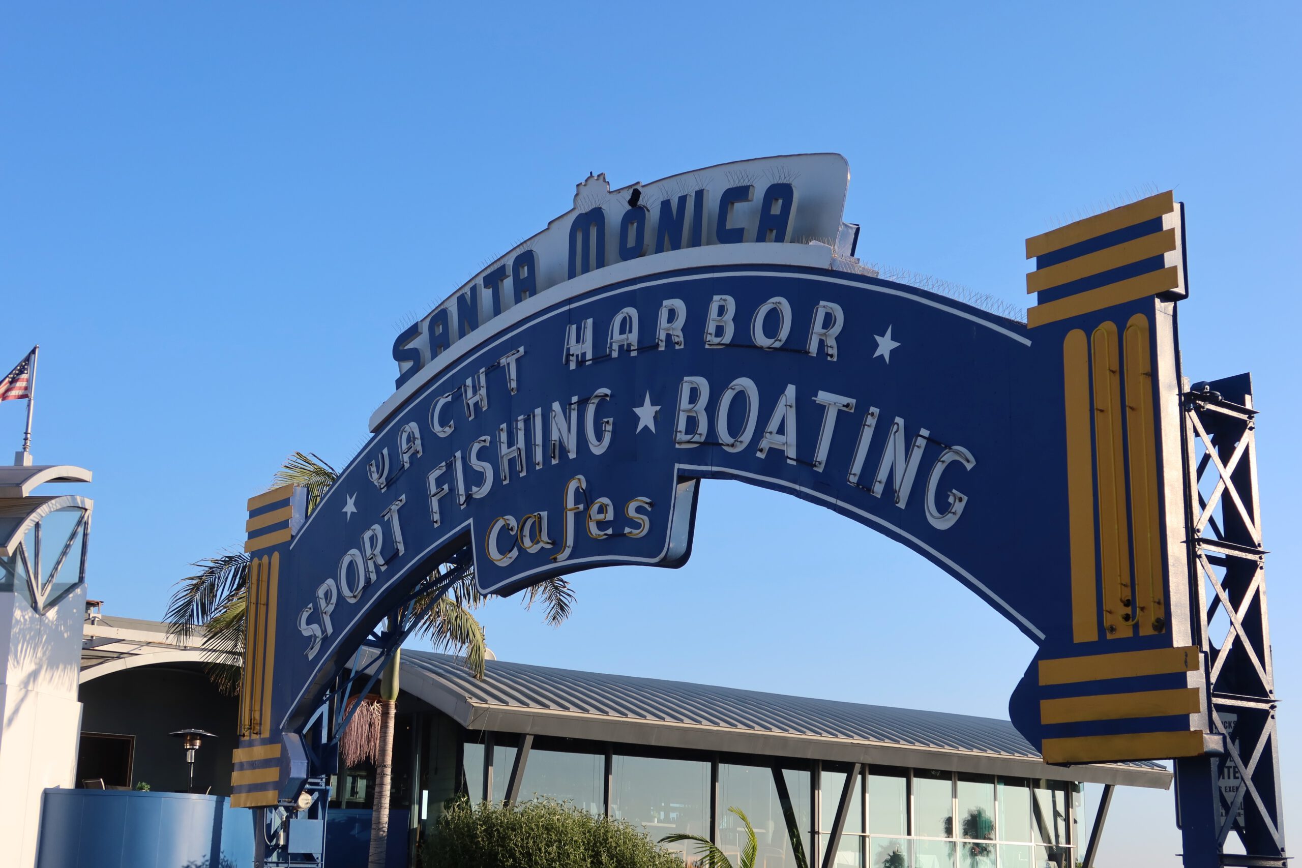 Iconic blue Santa Monica Pier entrance sign reading “Santa Monica Yacht Harbor, Sport Fishing, Boating, Cafes,” framed by palm trees and pier buildings, photographed under a clear blue California sky near the entrance to Santa Monica Pier.