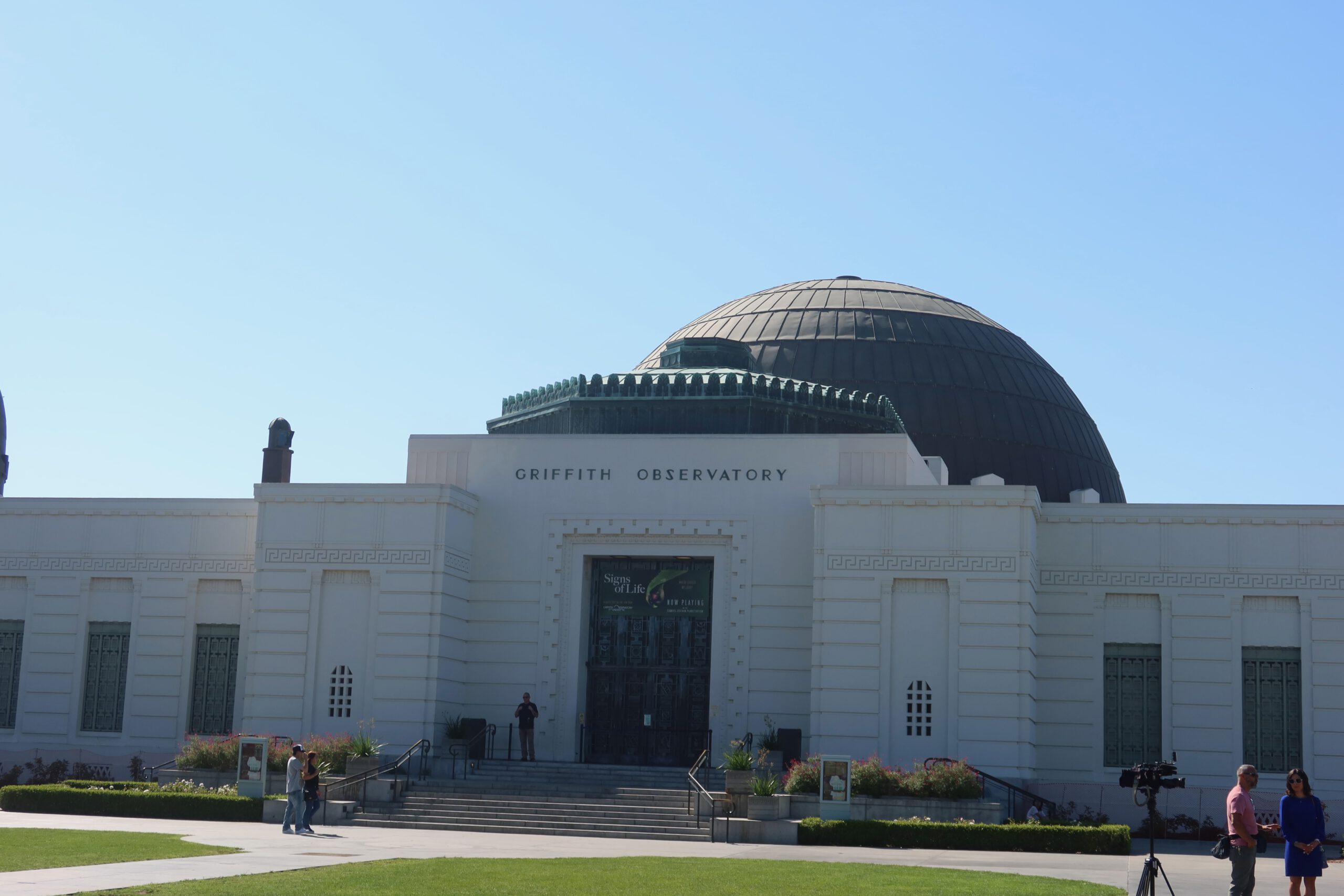 Front view of Griffith Observatory with its iconic white Art Deco façade and large central dome, visitors walking across the lawn under a clear blue sky in Los Angeles.