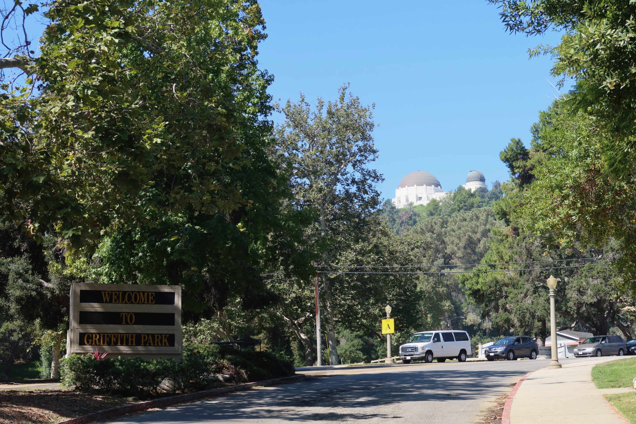 Entrance road to Griffith Park with the “Welcome to Griffith Park” sign in the foreground and Griffith Observatory visible on the hill in the distance under a clear blue sky in Los Angeles.