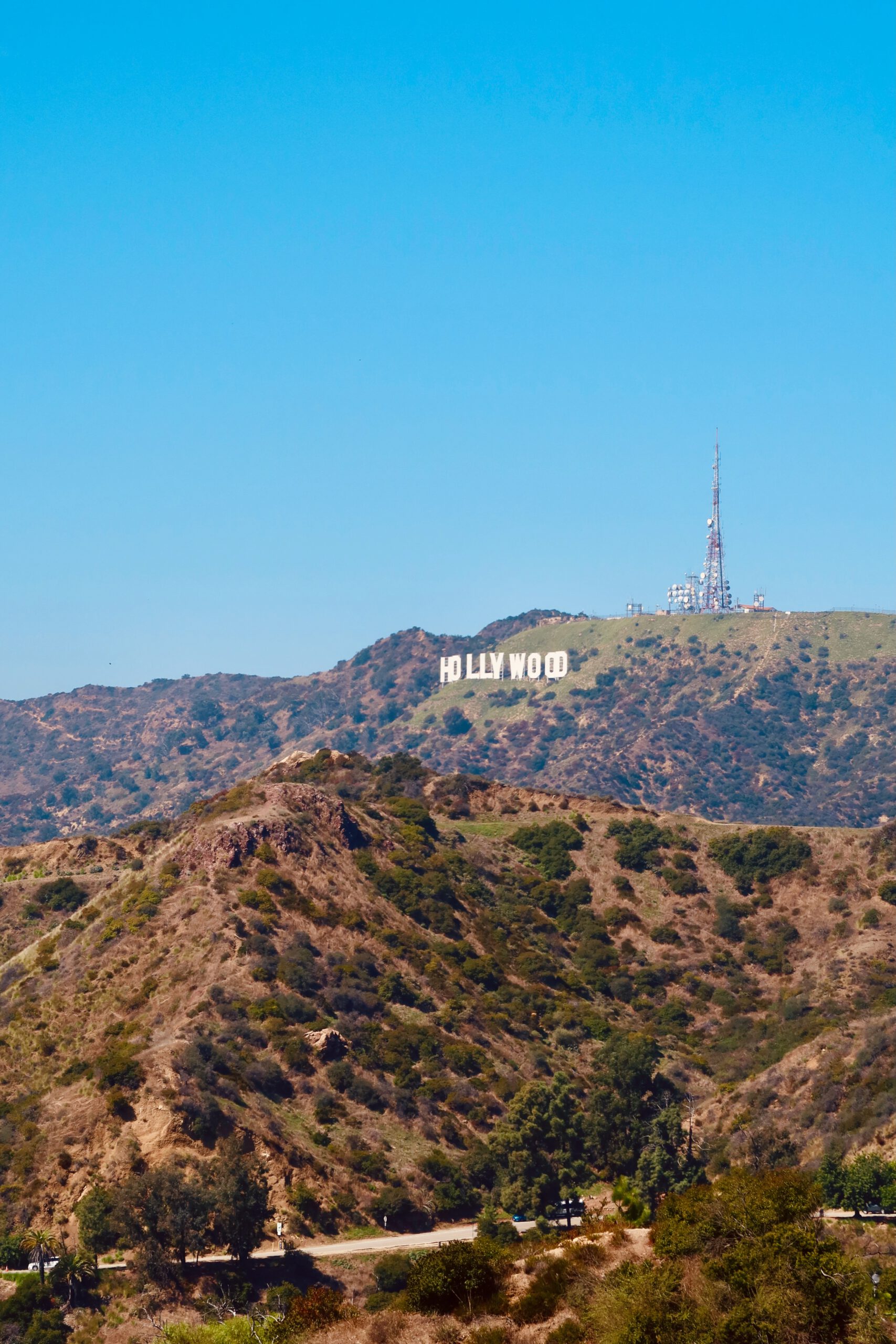Clear daytime view of the Hollywood Sign on the hillside, seen from Griffith Park, with rugged hills, dry vegetation, and communication towers under a bright blue sky in Los Angeles.