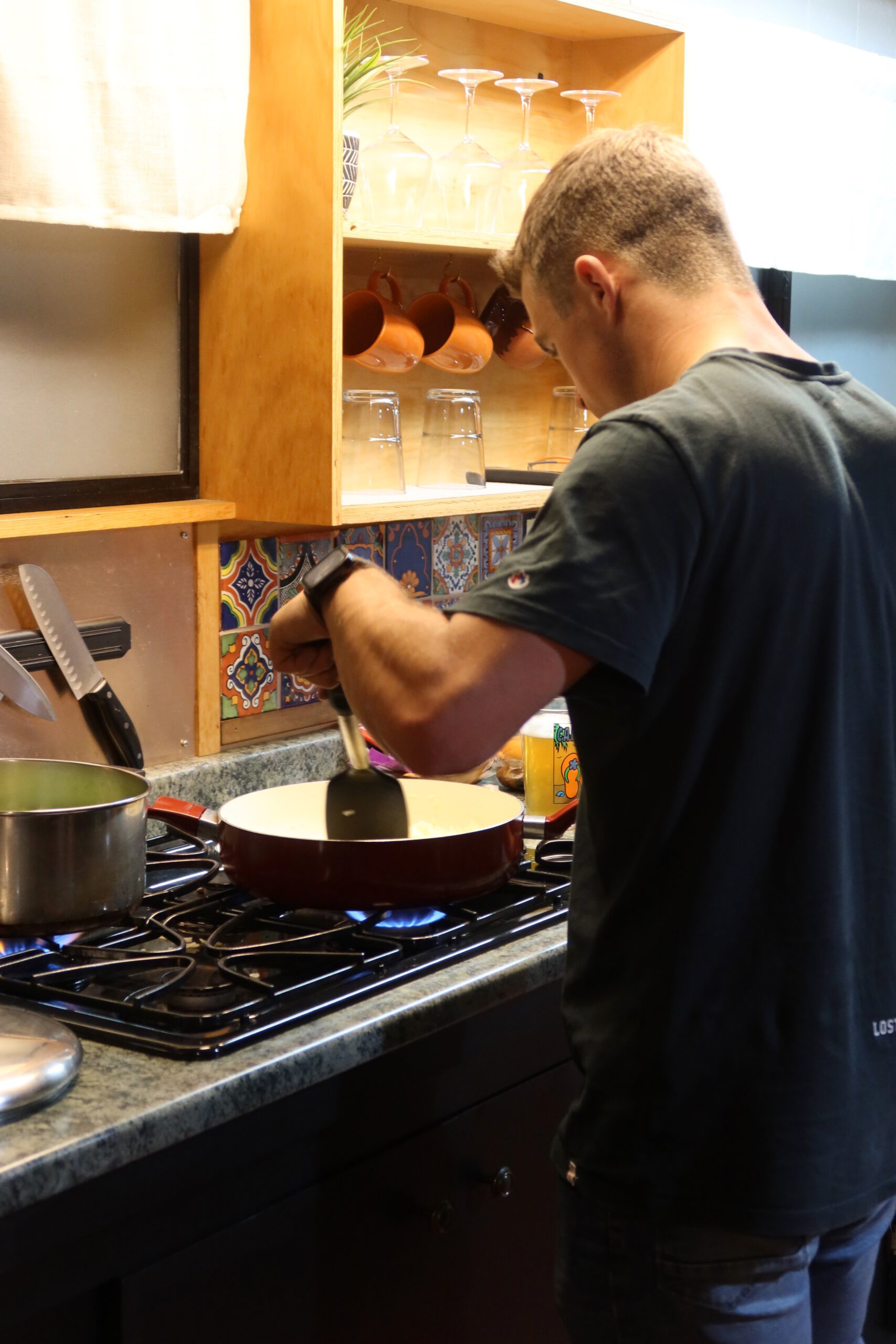 Cooking dinner in the kitchen at Casa Rustica Morongo Valley Airbnb, with a gas stove, frying pan on open flame, rustic shelves with glasses and mugs, and colorful tile backsplash.