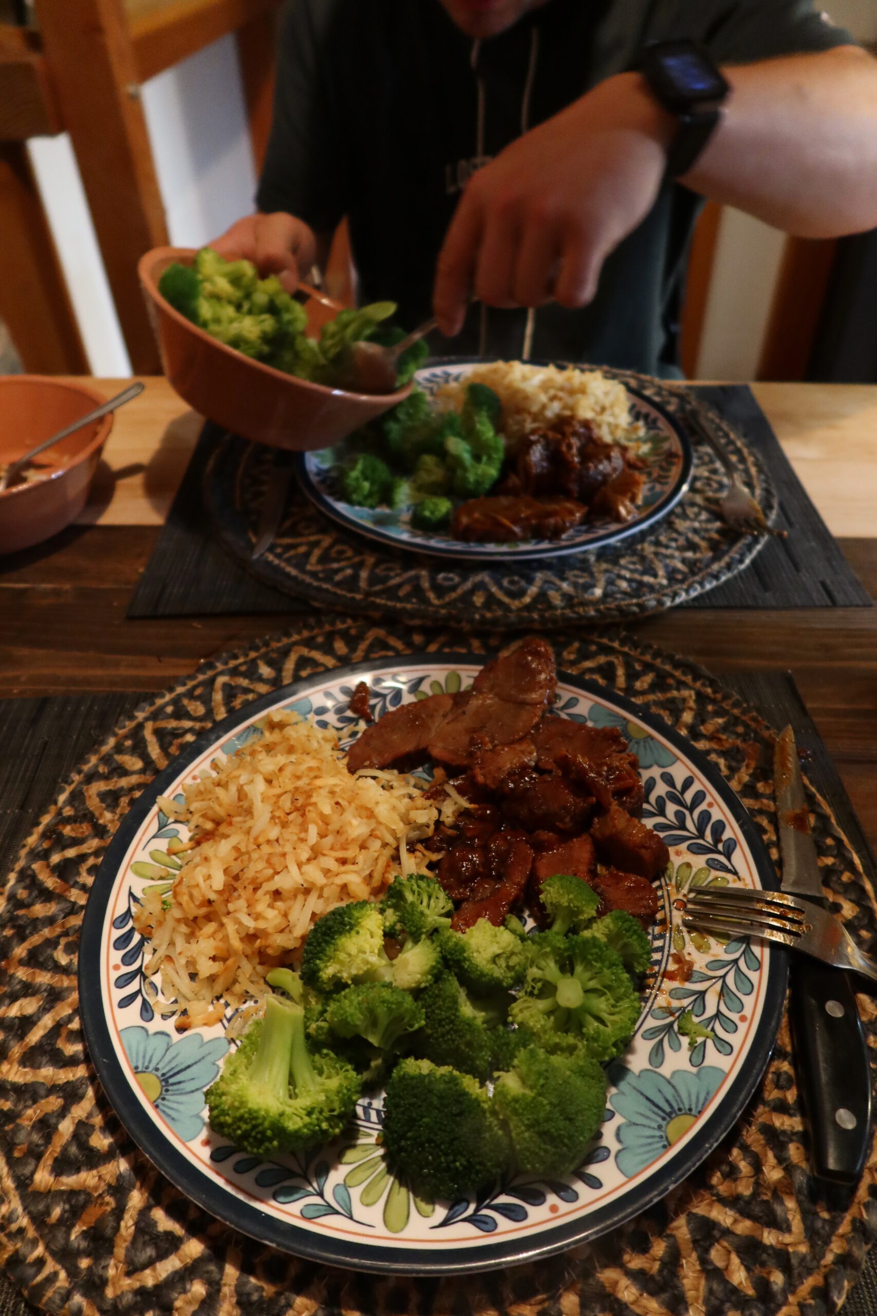 Home-cooked dinner at Casa Rustica Morongo Valley Airbnb featuring rice, broccoli, and cooked meat served on patterned plates at a wooden dining table, capturing a cozy evening meal.