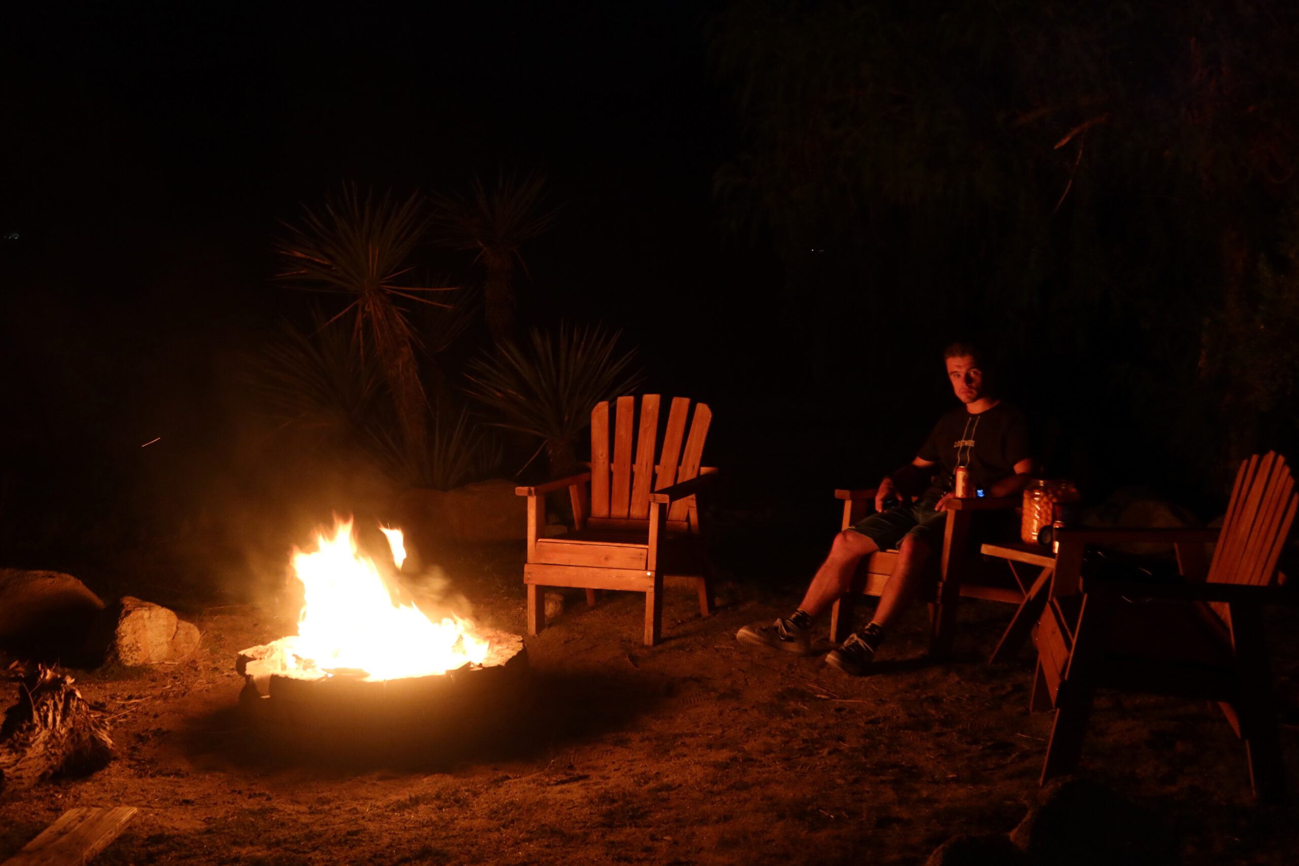 Evening fire pit at Casa Rustica Morongo Valley Airbnb with wooden outdoor chairs, warm firelight, desert plants in the background, and a quiet night atmosphere in Morongo Valley.
