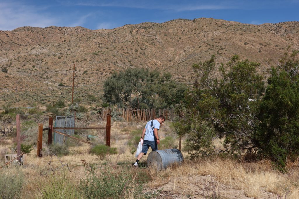 Person walking across the desert grounds at Casa Rustica Morongo Valley Airbnb, surrounded by dry grass, wooden fencing, shrubs, and mountain hills in the background under a clear blue sky.
