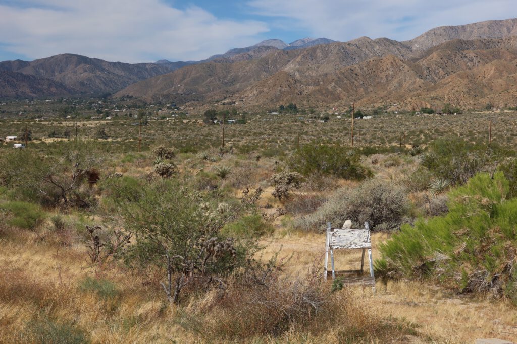 Wide view of the Morongo Valley desert landscape with scattered shrubs, dry vegetation, distant houses, and rugged mountain ranges stretching across the horizon.