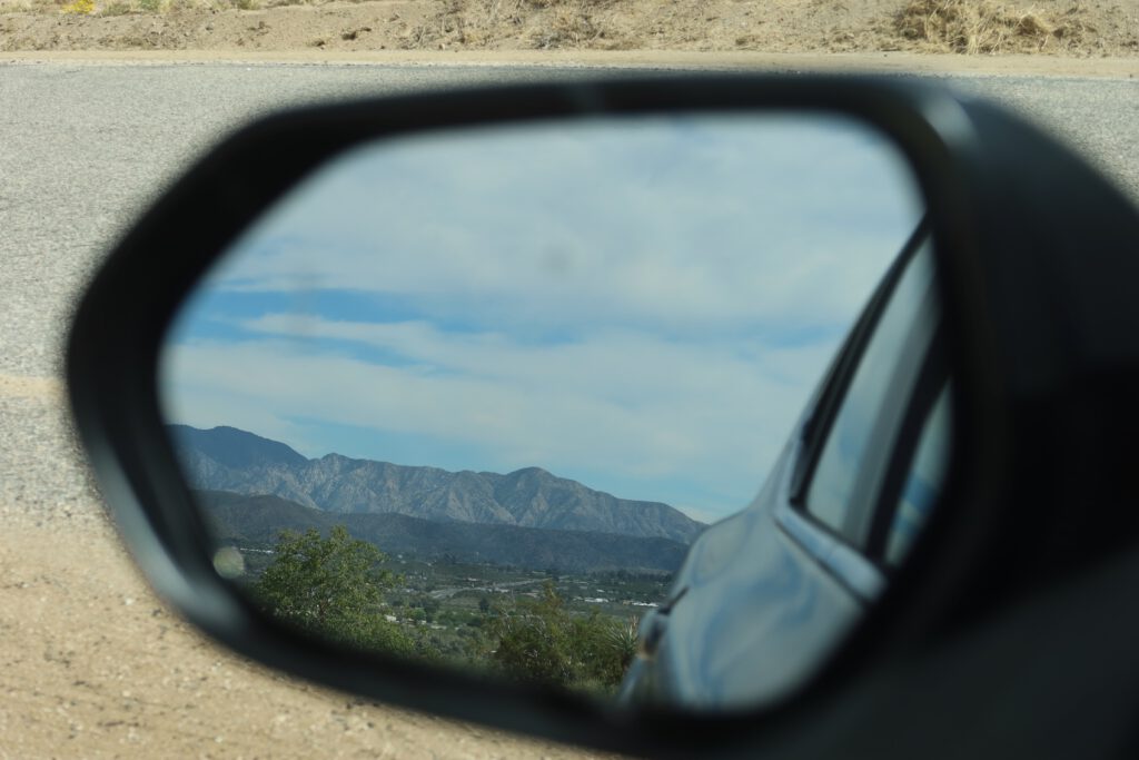 Mountain landscape reflected in a car side mirror along a desert road near Morongo Valley, capturing the rugged hills and open sky from a road trip perspective.