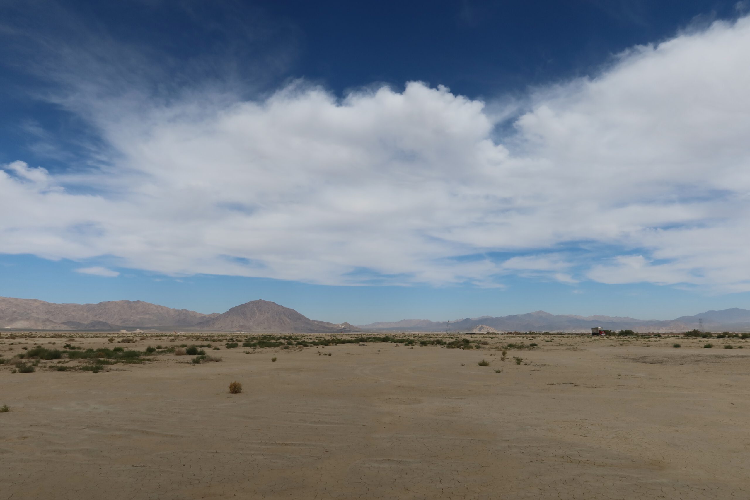 Mojave Desert California landscape with wide sandy plains, sparse desert vegetation, and distant mountain ranges under dramatic cloud formations.