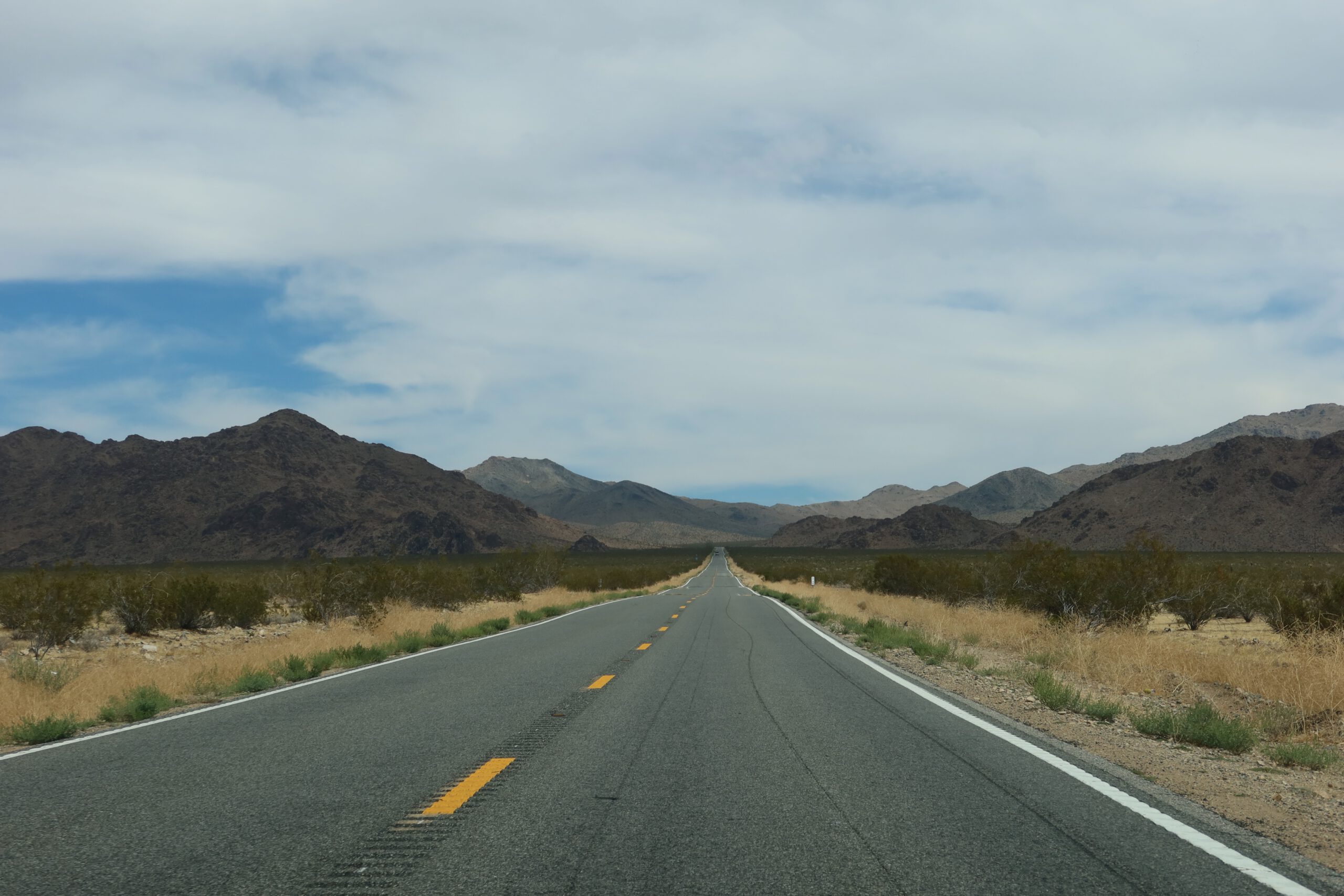 Mojave Desert road trip view of a straight desert highway cutting through a vast Mojave Desert landscape, framed by rugged mountains and dry grasslands.