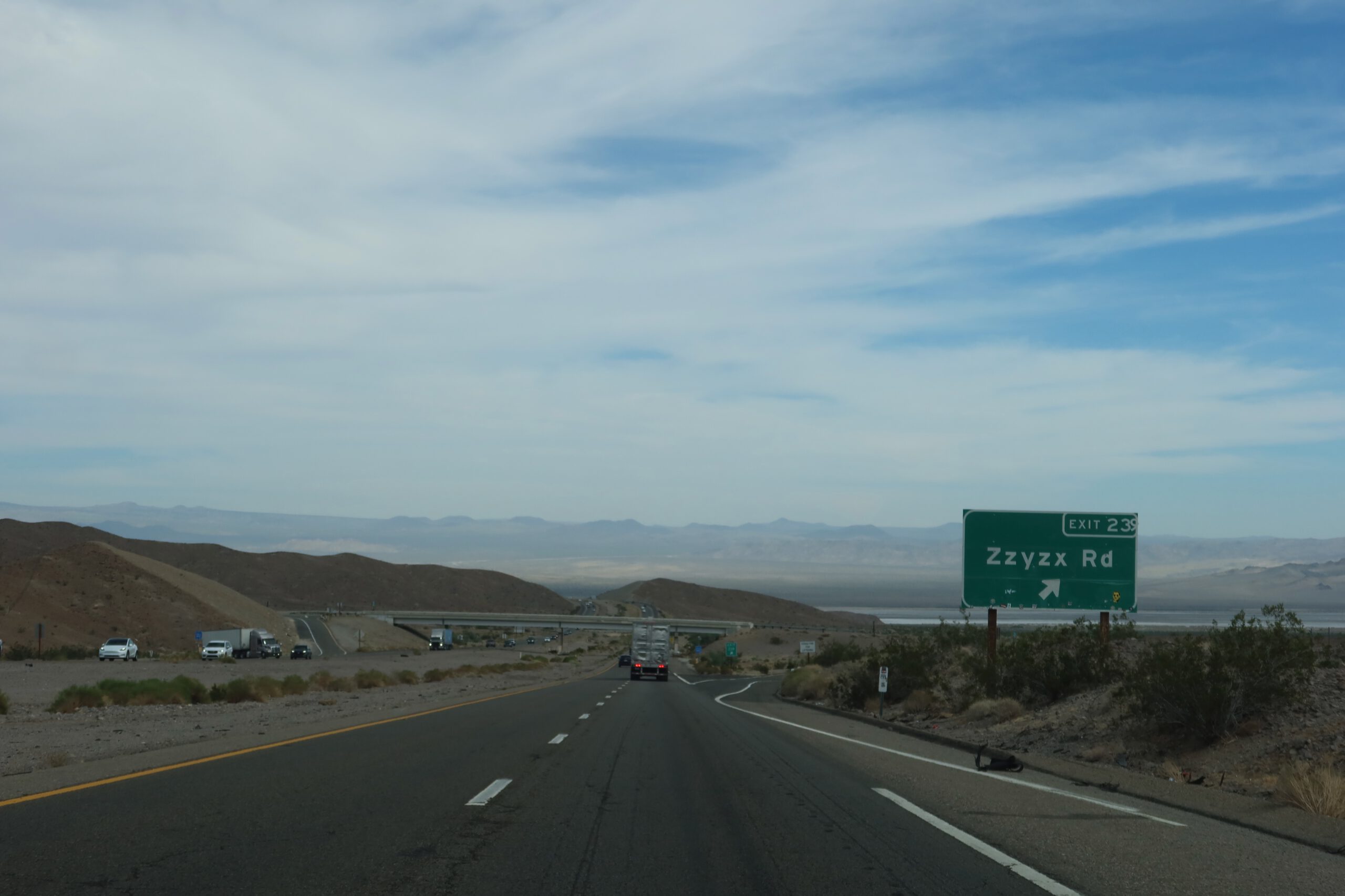 Mojave Desert road trip on a California highway approaching the Zzyzx Road exit, with open desert plains, surrounding hills, and expansive Mojave Desert landscape.