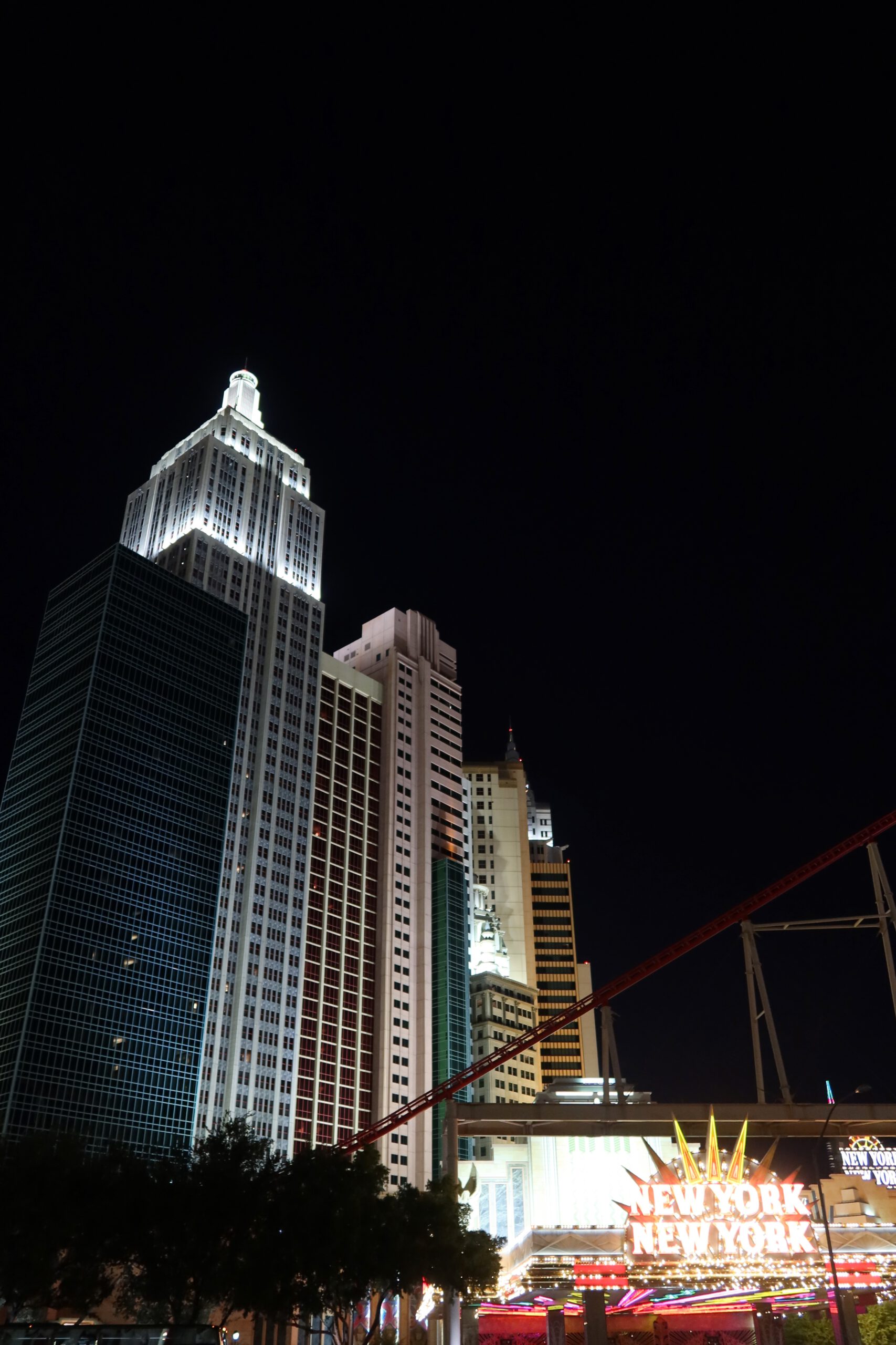 Night view of the New York-New York Hotel & Casino skyline in Las Vegas, featuring illuminated skyscraper replicas and the red roller coaster track against a dark sky.