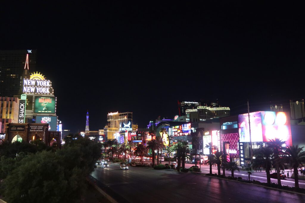 Wide nighttime view of the Las Vegas Strip with New York-New York, neon-lit hotels, palm trees, and busy traffic along Las Vegas Boulevard.