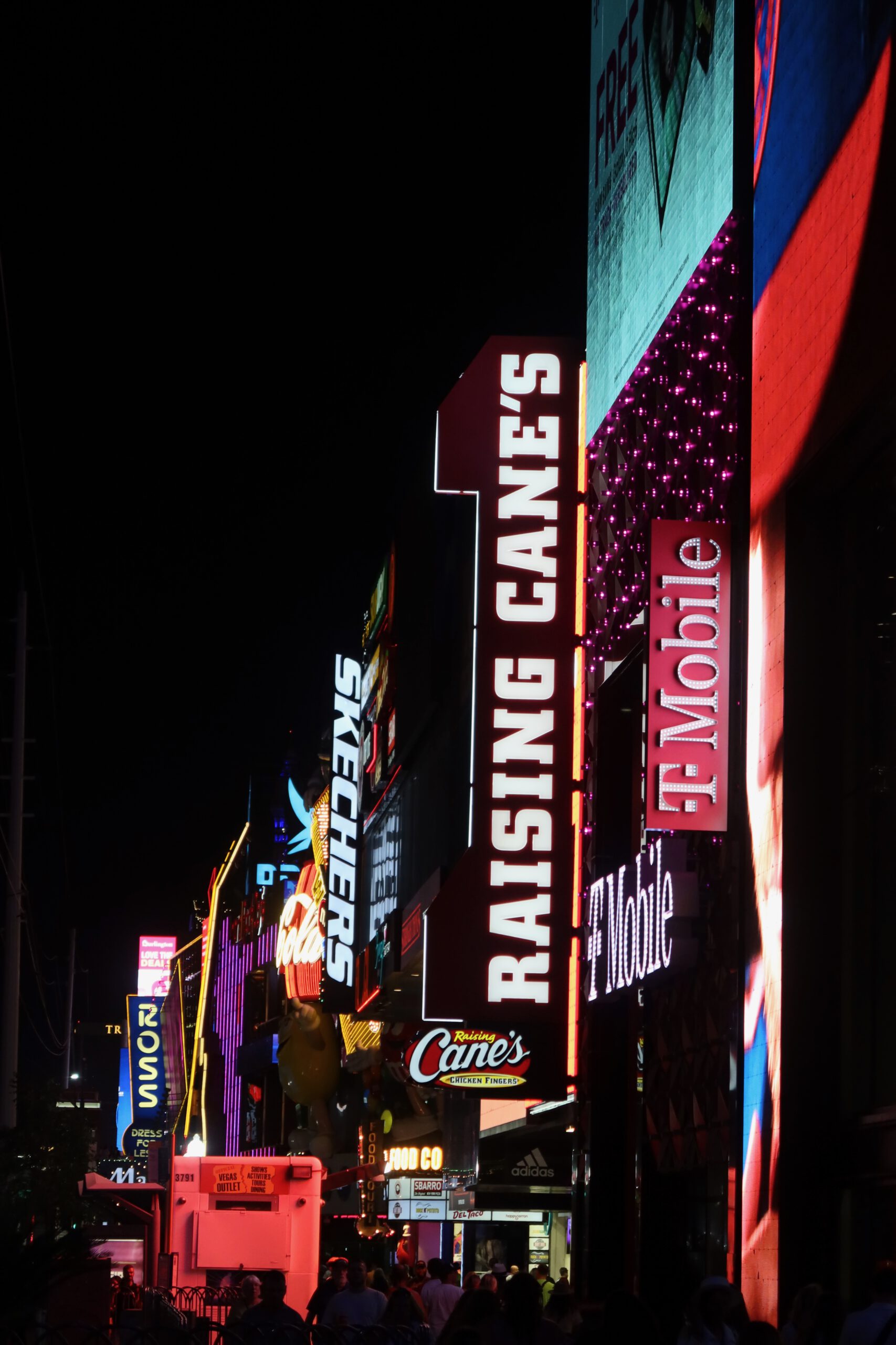 Vertical nighttime view of the Las Vegas Strip with bright neon signs for Raising Cane’s, T-Mobile, Skechers, and other storefronts, crowded sidewalks, and glowing billboards.