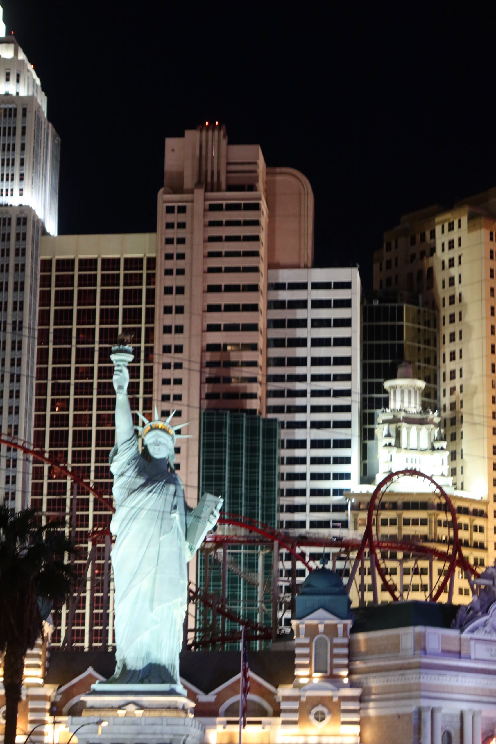 Replica of the Statue of Liberty at New York-New York Hotel & Casino in Las Vegas, lit at night with surrounding skyscraper facades and roller coaster tracks.