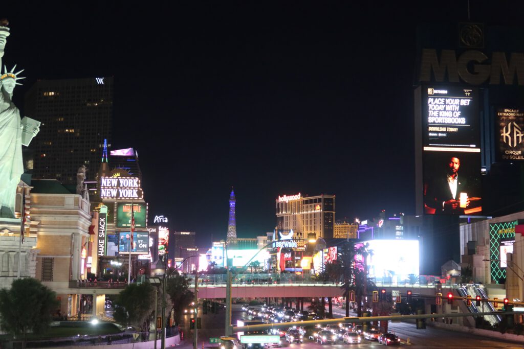 Night view of the Las Vegas Strip featuring New York-New York, MGM Grand signage, bright digital billboards, and heavy traffic below.