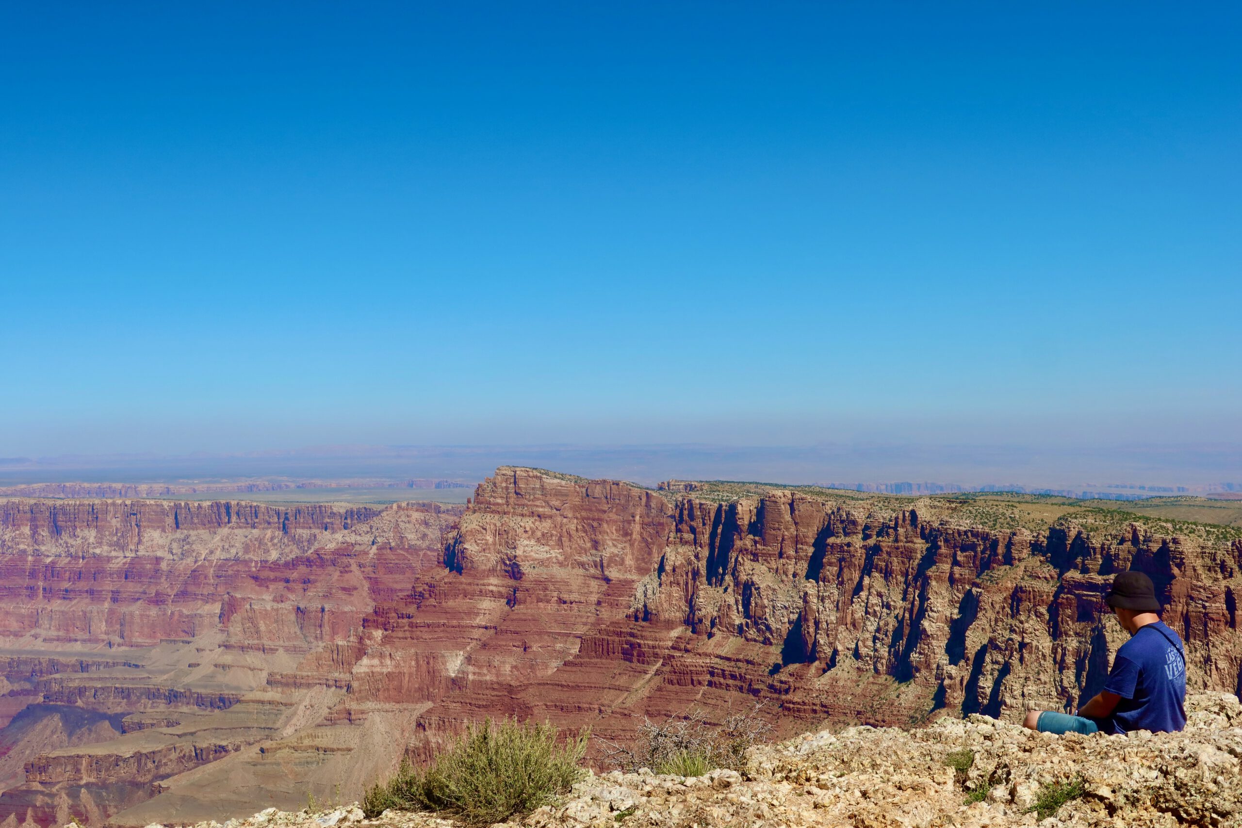 Traveler sitting at the edge of the Grand Canyon, taking in the vast canyon landscape under a clear blue sky