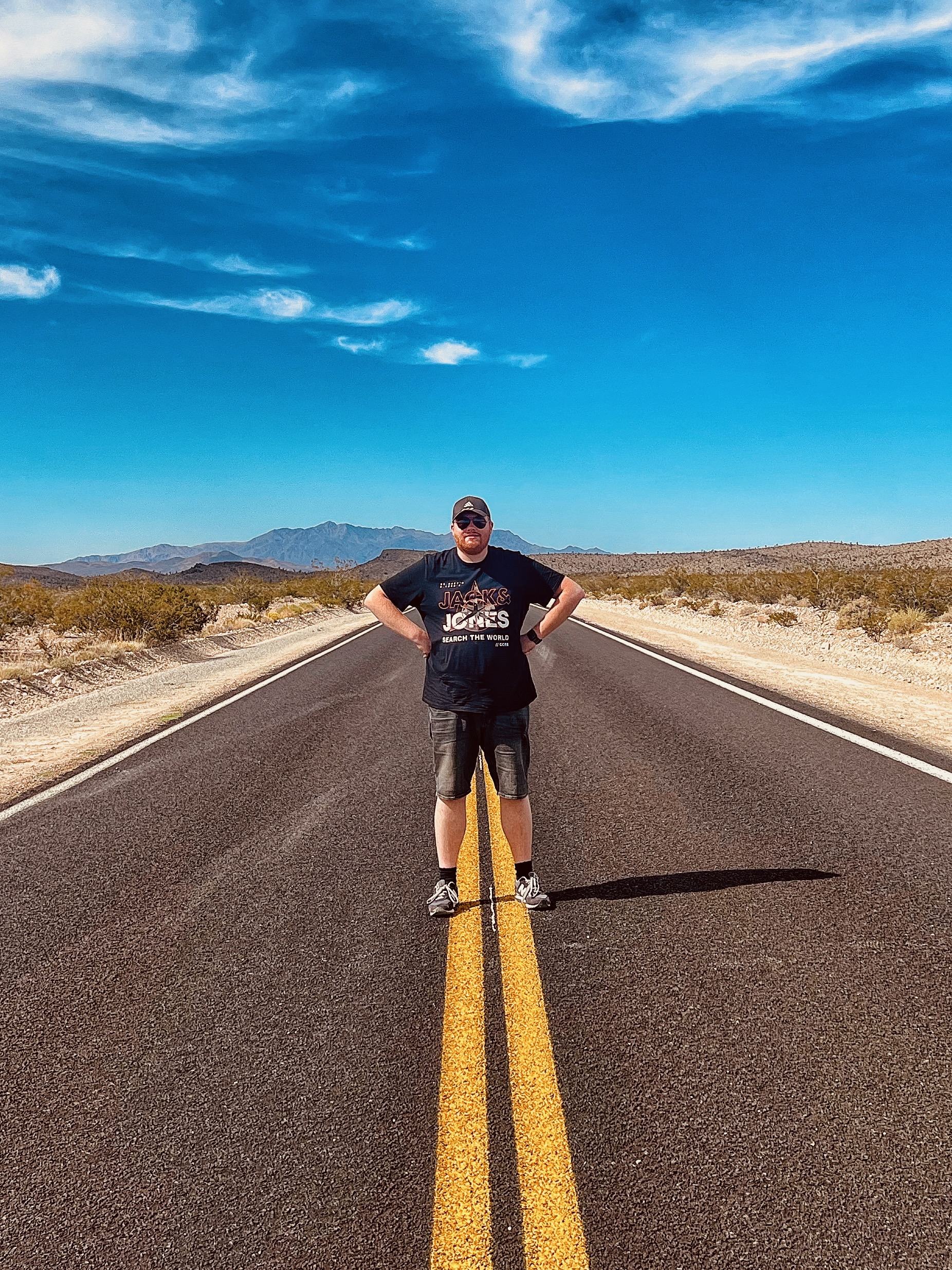 Mojave Desert road trip moment in California with a person standing on the center line of an empty desert highway, surrounded by wide Mojave Desert landscape and distant mountain ranges under a bright blue sky.
