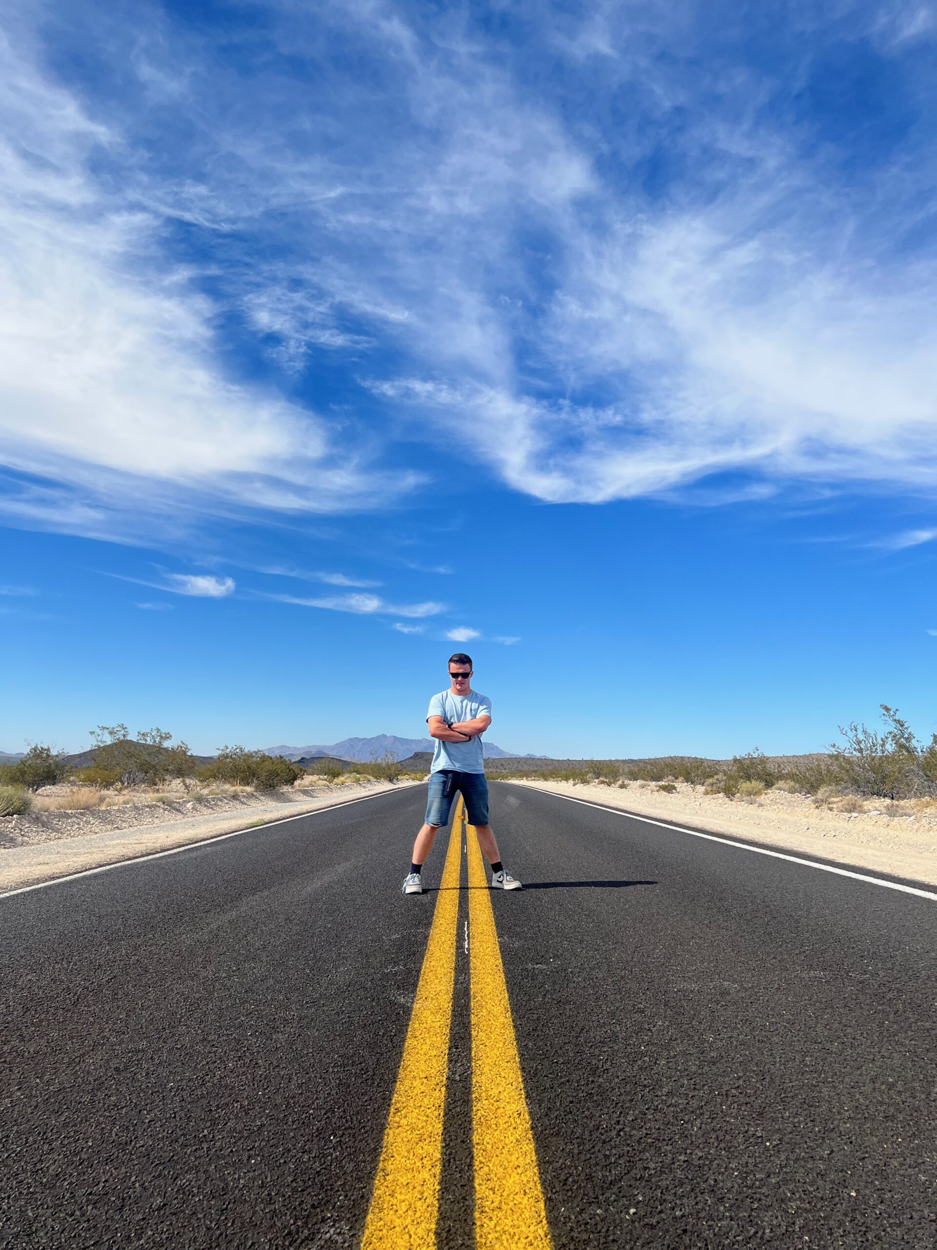 Driving through the Mojave Desert in California with a person standing on a straight desert road, yellow center lines leading into an open Mojave Desert landscape with clear skies and desert vegetation.