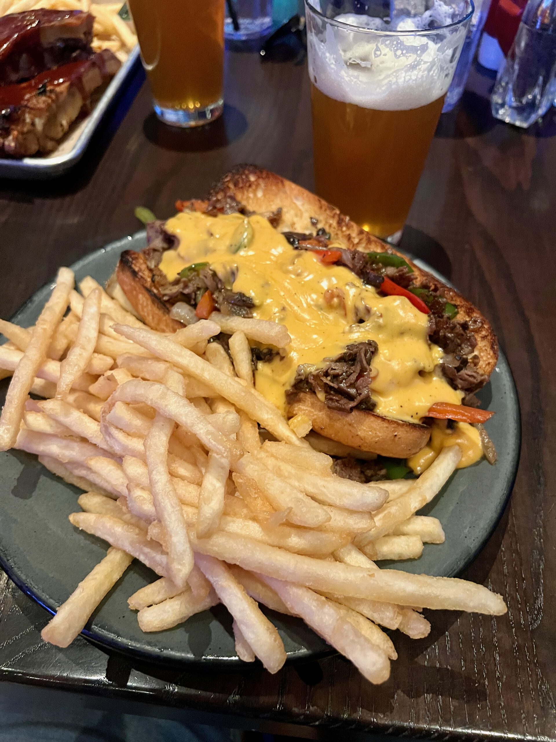 Loaded steak sandwich with melted cheese, grilled vegetables, and French fries served at Public House in Luxor Hotel & Casino Las Vegas, with two glasses of draft beer on the table.