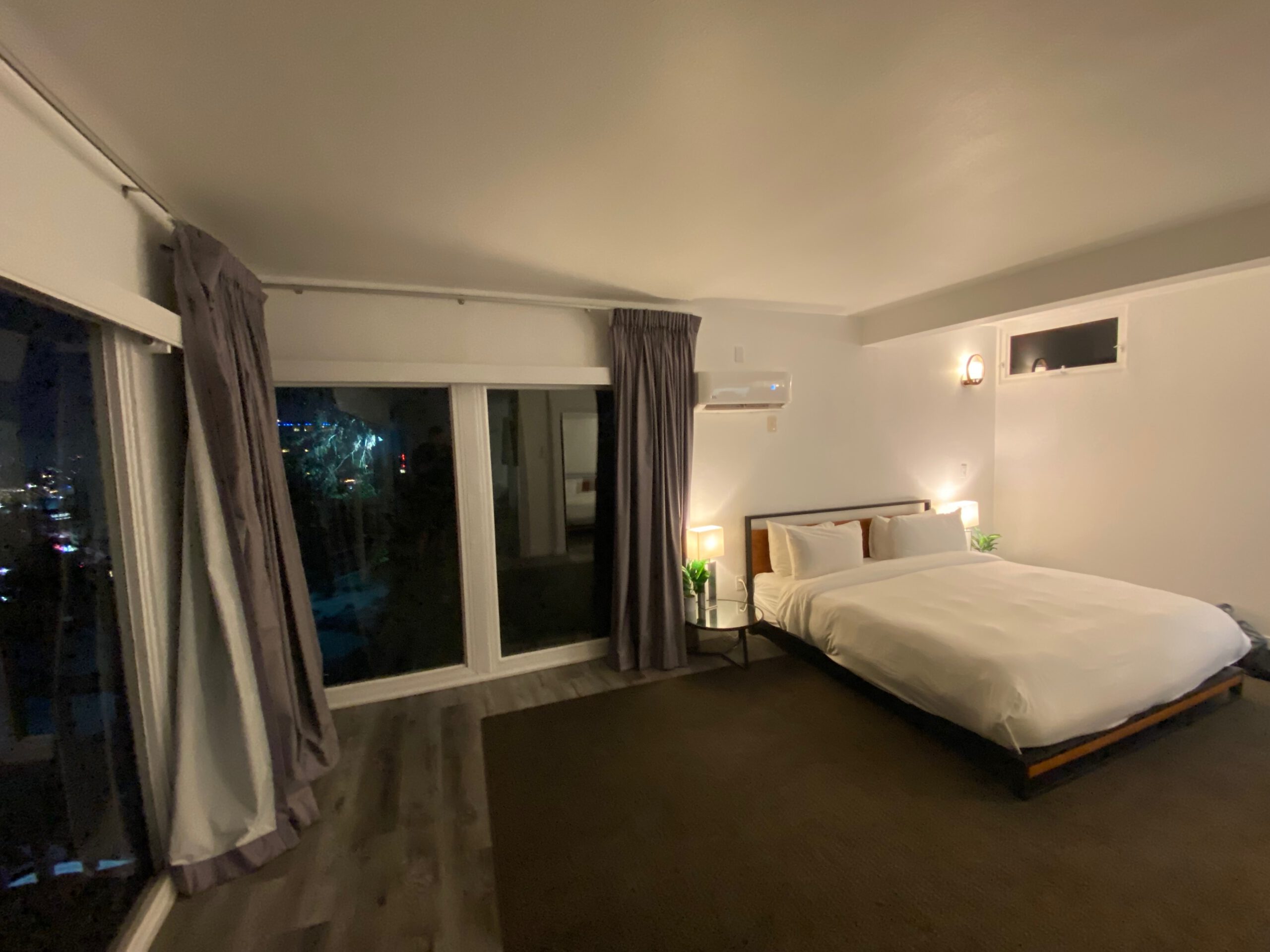Spacious hotel room in the Hollywood Hills with a large bed, floor-to-ceiling windows, curtains, and nighttime city lights visible outside.