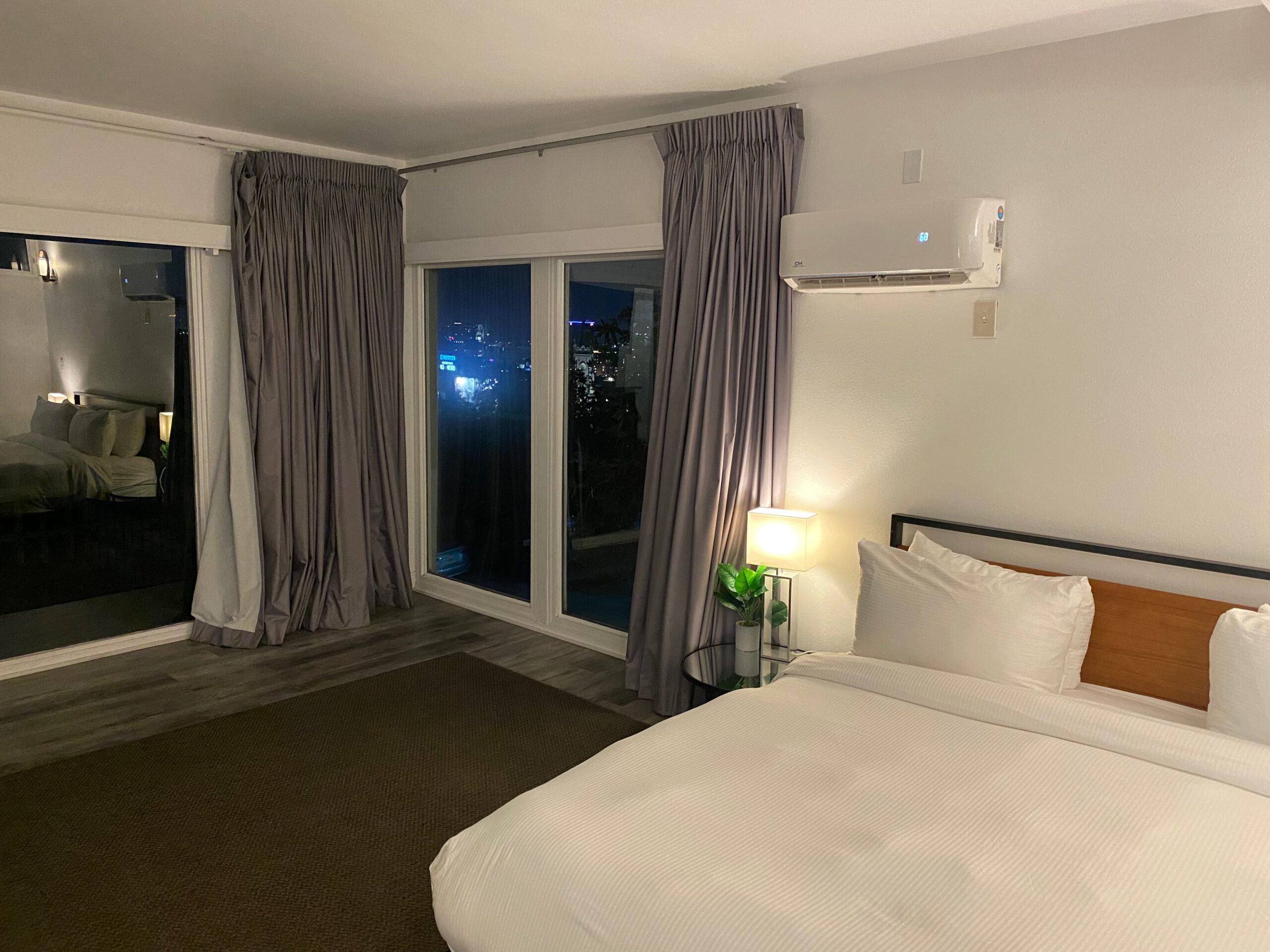 Modern hotel bedroom in the Hollywood Hills featuring a large bed, bedside lighting, air conditioning unit, and window overlooking Los Angeles at night.