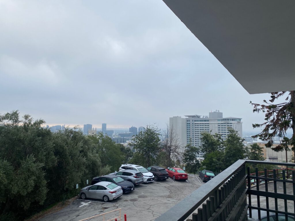 View from a Hollywood Hills hotel overlooking a small parking area with cars, trees, and Los Angeles skyline in the distance under an overcast sky.