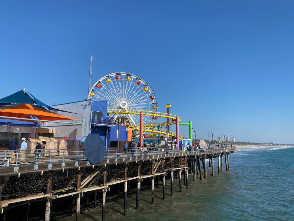 Side view of Santa Monica Pier with Pacific Park attractions lining the wooden pier, including the Ferris wheel and yellow roller coaster, overlooking the Pacific Ocean with beach and coastline stretching into the distance under bright blue skies.