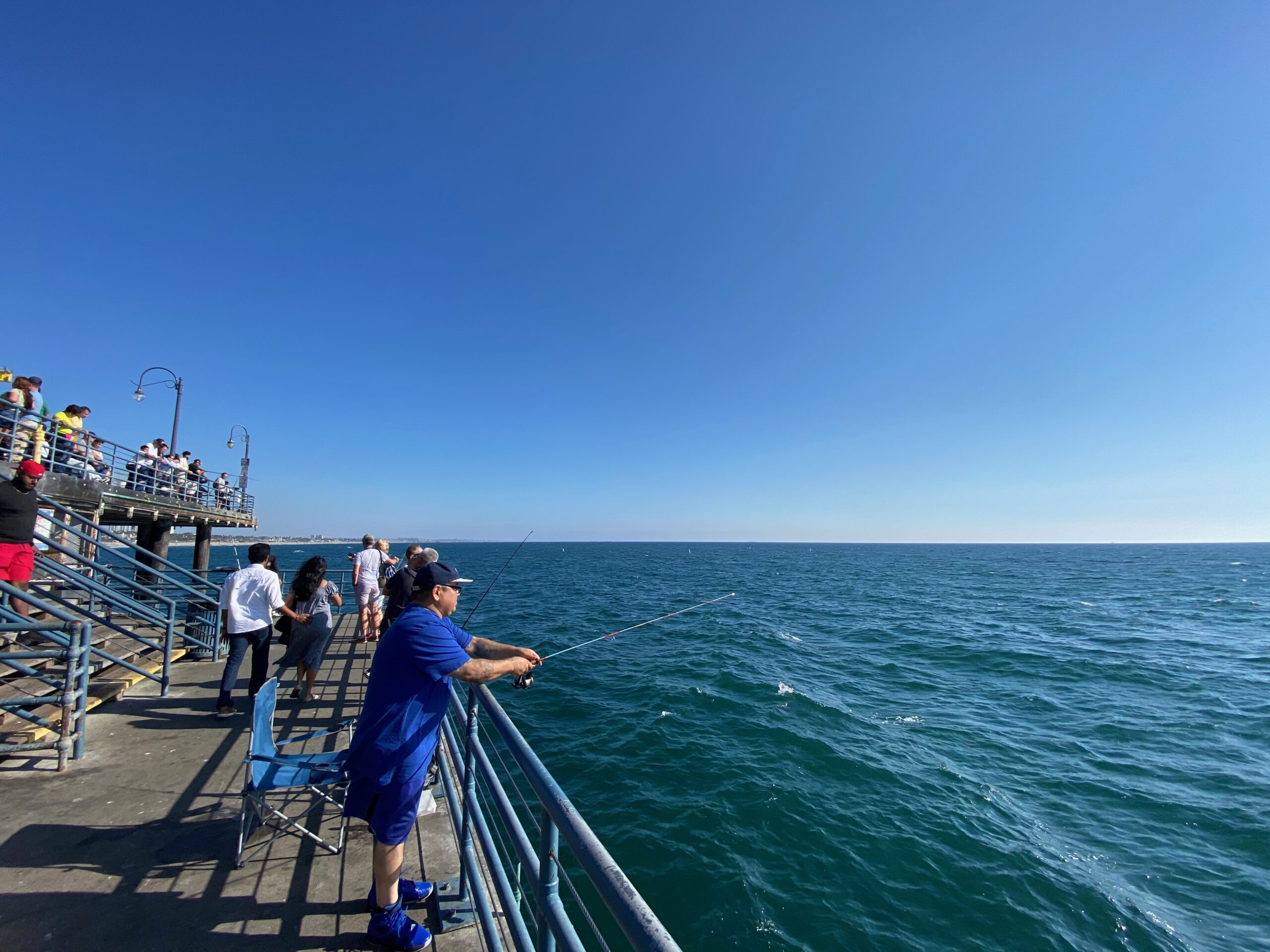 People fishing from the railing of Santa Monica Pier, looking out over the Pacific Ocean with fishing rods extended above clear blue water on a bright afternoon.