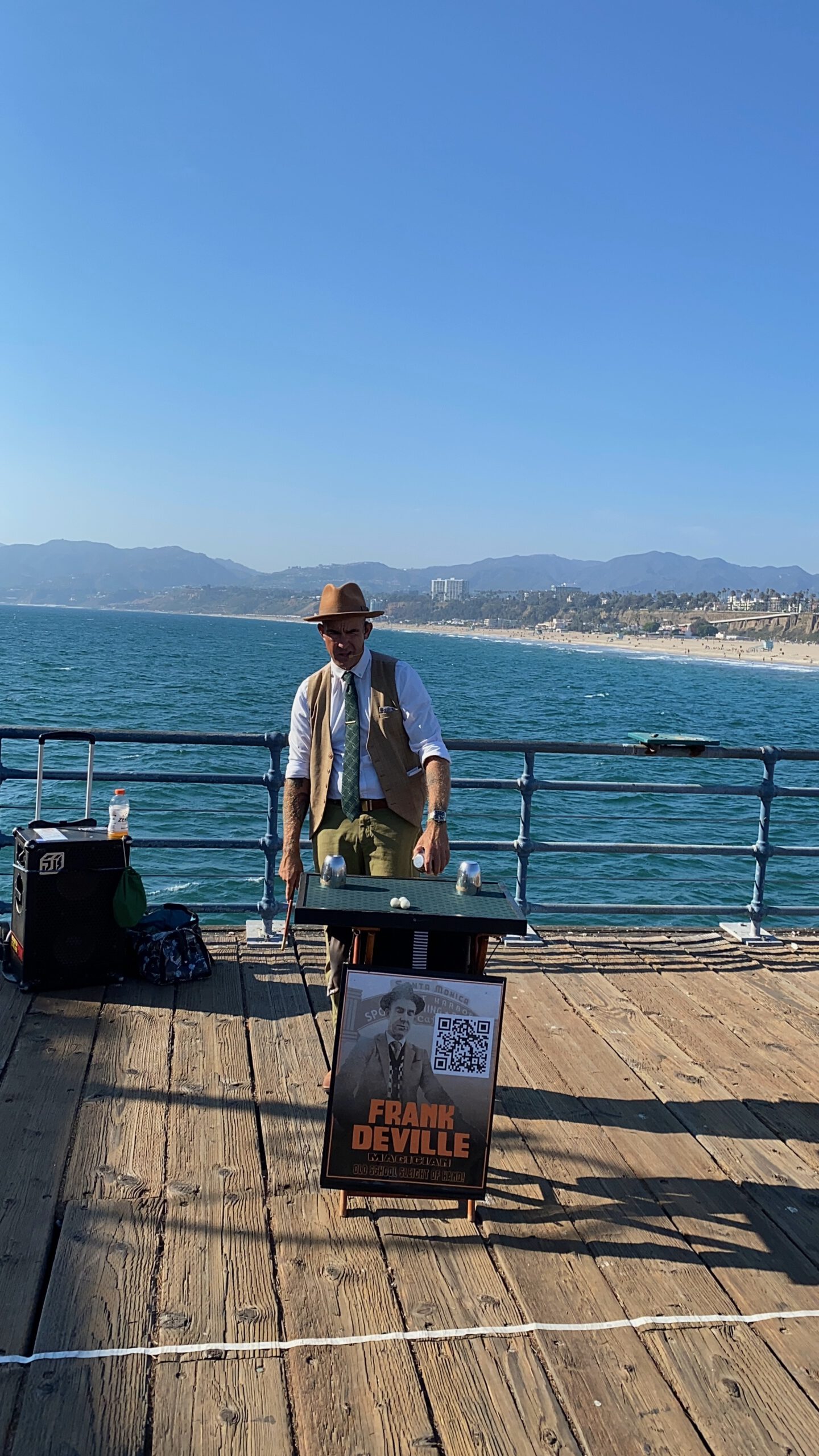 Street magician Frank Deville performing a classic cups-and-balls magic routine on Santa Monica Pier, standing on the wooden pier deck with the Pacific Ocean behind him, distant California coastline and mountains in the background, under a clear blue sky.