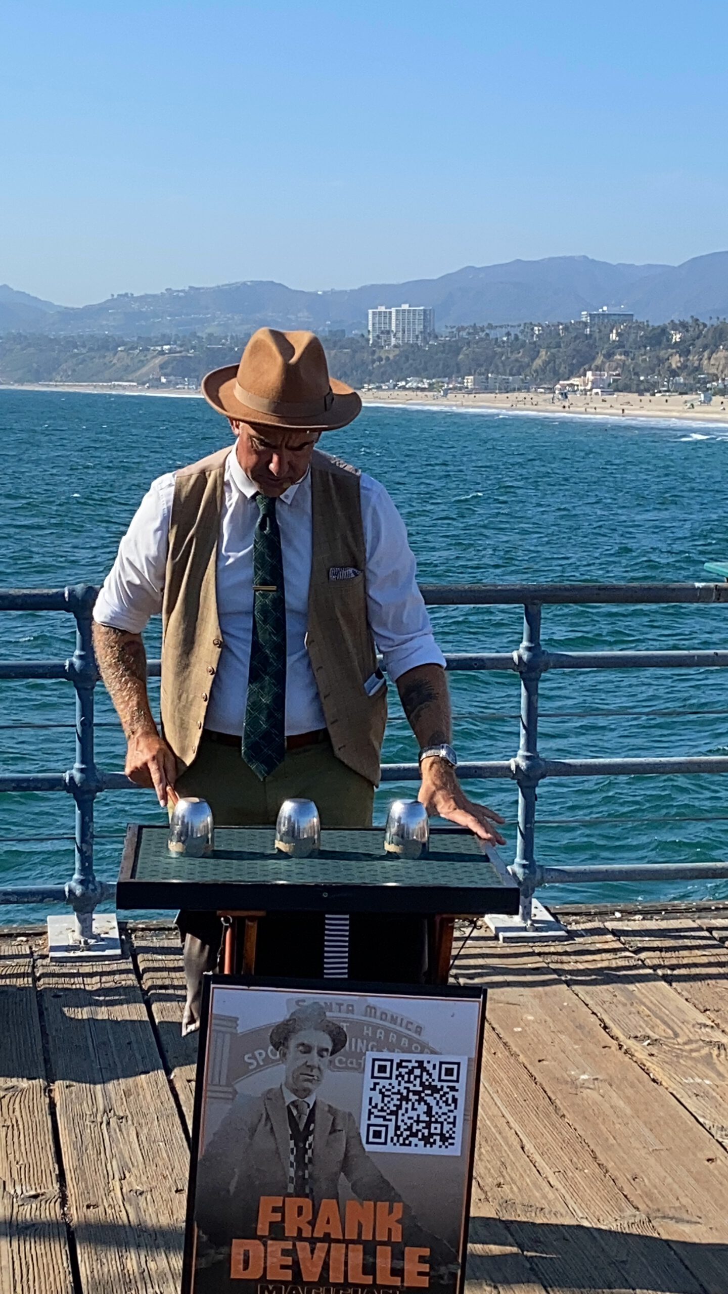 Close-up view of street magician Frank Deville at Santa Monica Pier, wearing a vintage-style outfit with hat, vest, and tie while demonstrating a cups-and-balls magic trick, with the ocean railing, blue water, and Santa Monica coastline visible behind him.