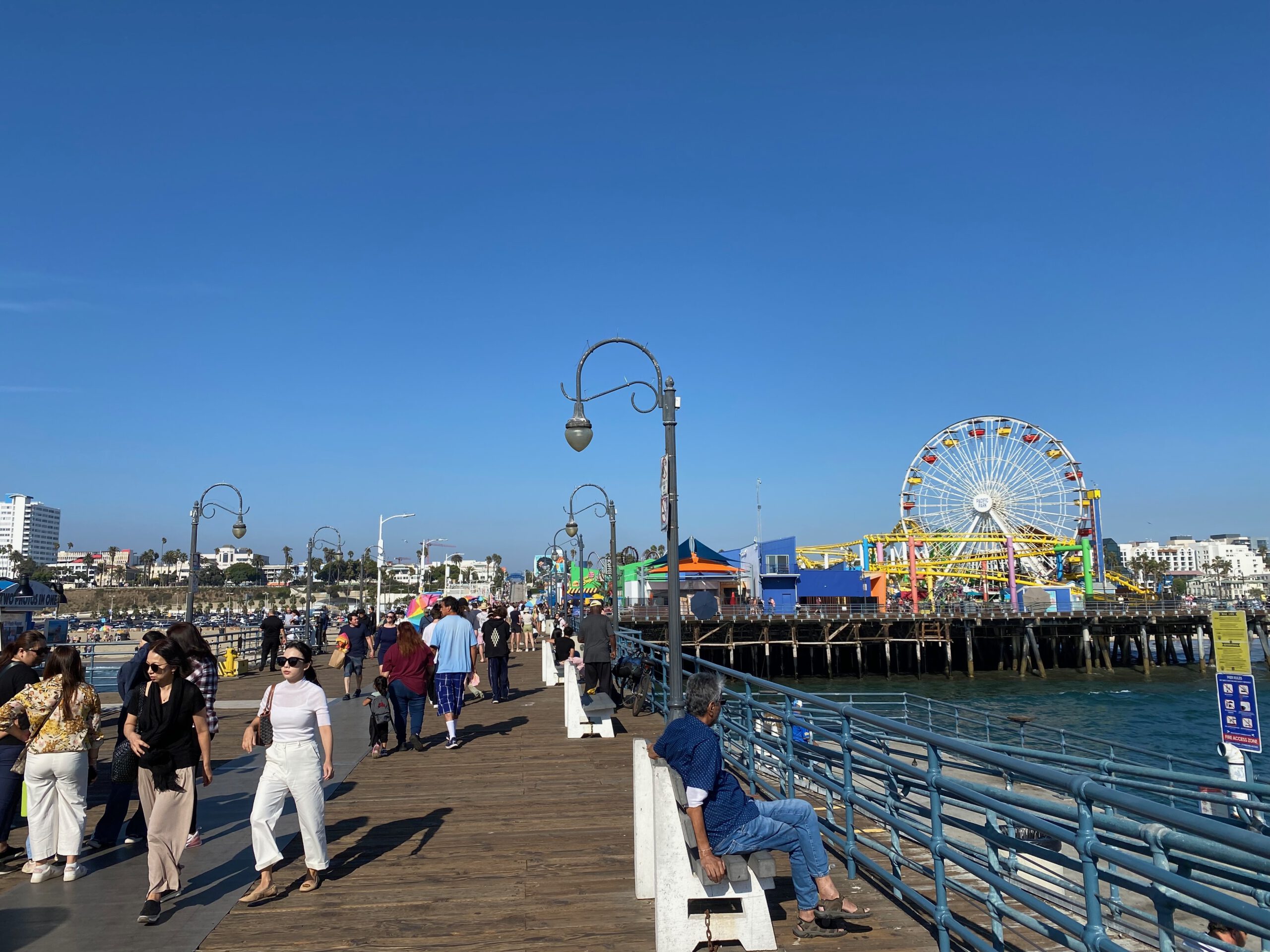 Crowds strolling along the wooden Santa Monica Pier with classic lampposts, ocean views, and Pacific Park’s Ferris wheel and roller coaster visible in the distance.