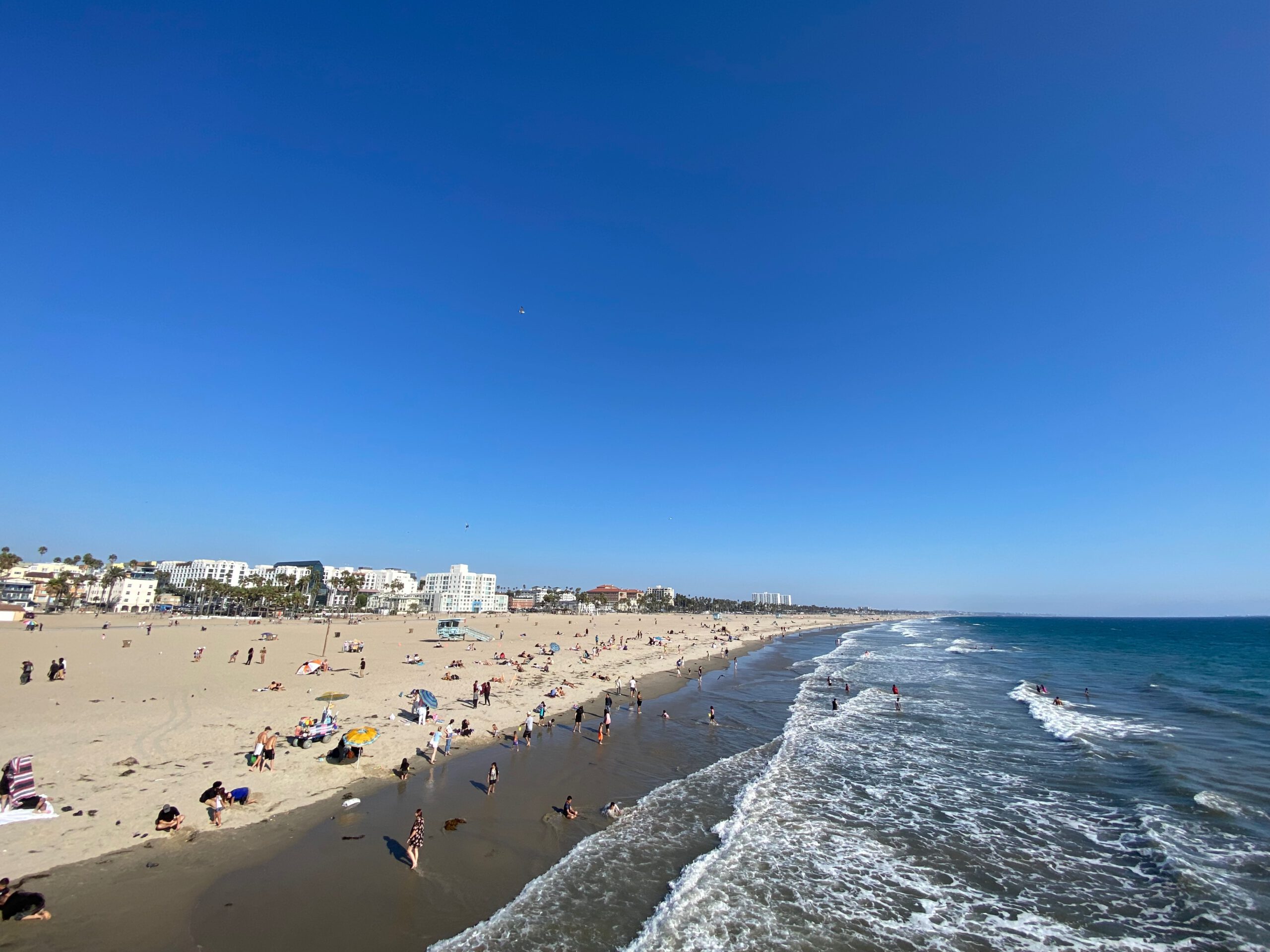 Wide panoramic view from Santa Monica Pier showing a long sandy beach filled with sunbathers and swimmers, gentle Pacific Ocean waves rolling along the shoreline, beach umbrellas and people scattered across the sand, and the Santa Monica coastline with palm trees and low-rise buildings under a clear deep-blue California sky.