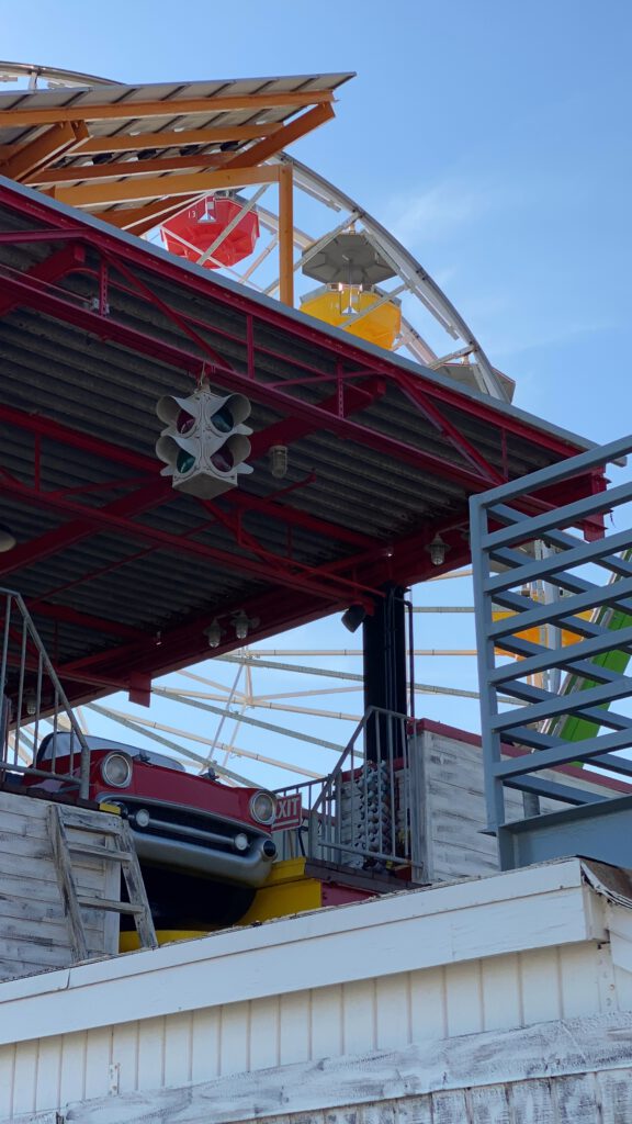 Detail view of a classic ride car emerging from a iron structure at Pacific Park on Santa Monica Pier, with colorful Ferris wheel gondolas visible above the roofline against a clear blue California sky.