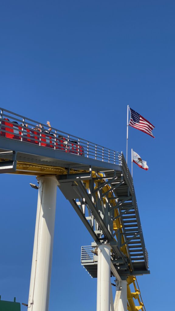 Roller coaster track at Pacific Park on Santa Monica Pier with riders raising their hands, framed by the American flag and California state flag waving beside the elevated track under a bright, cloudless blue sky.