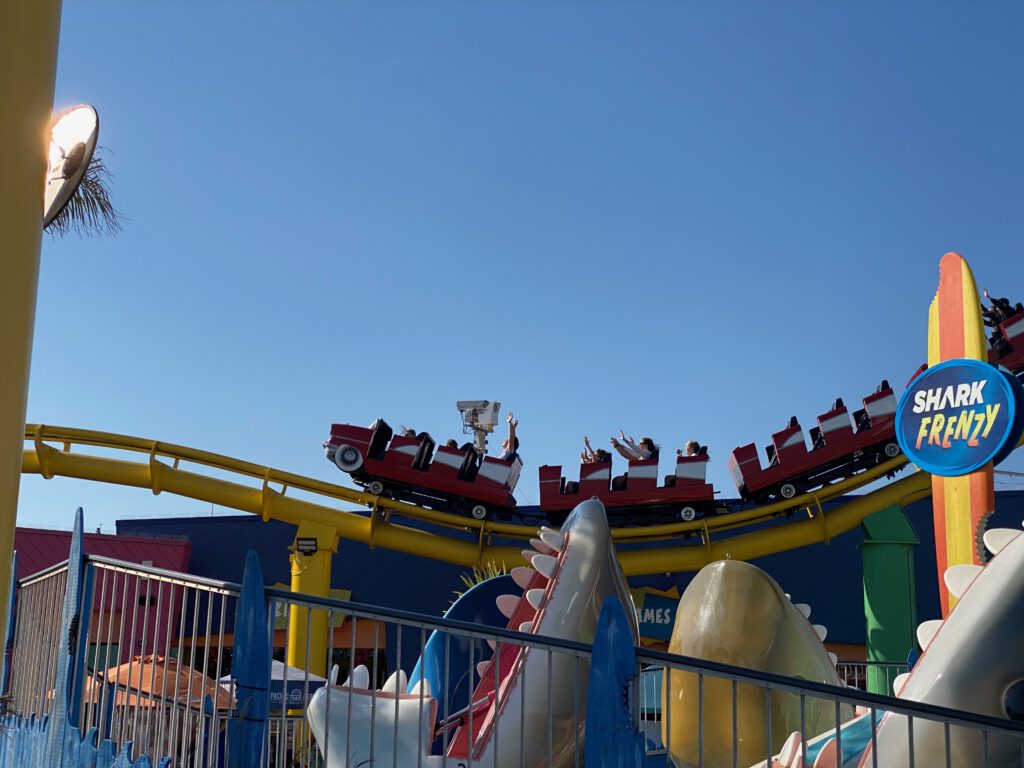 West Coaster roller coaster at Pacific Park on Santa Monica Pier, showing red ride cars filled with excited riders above shark-themed decorations, captured against a clear blue sky and vibrant amusement park colors.