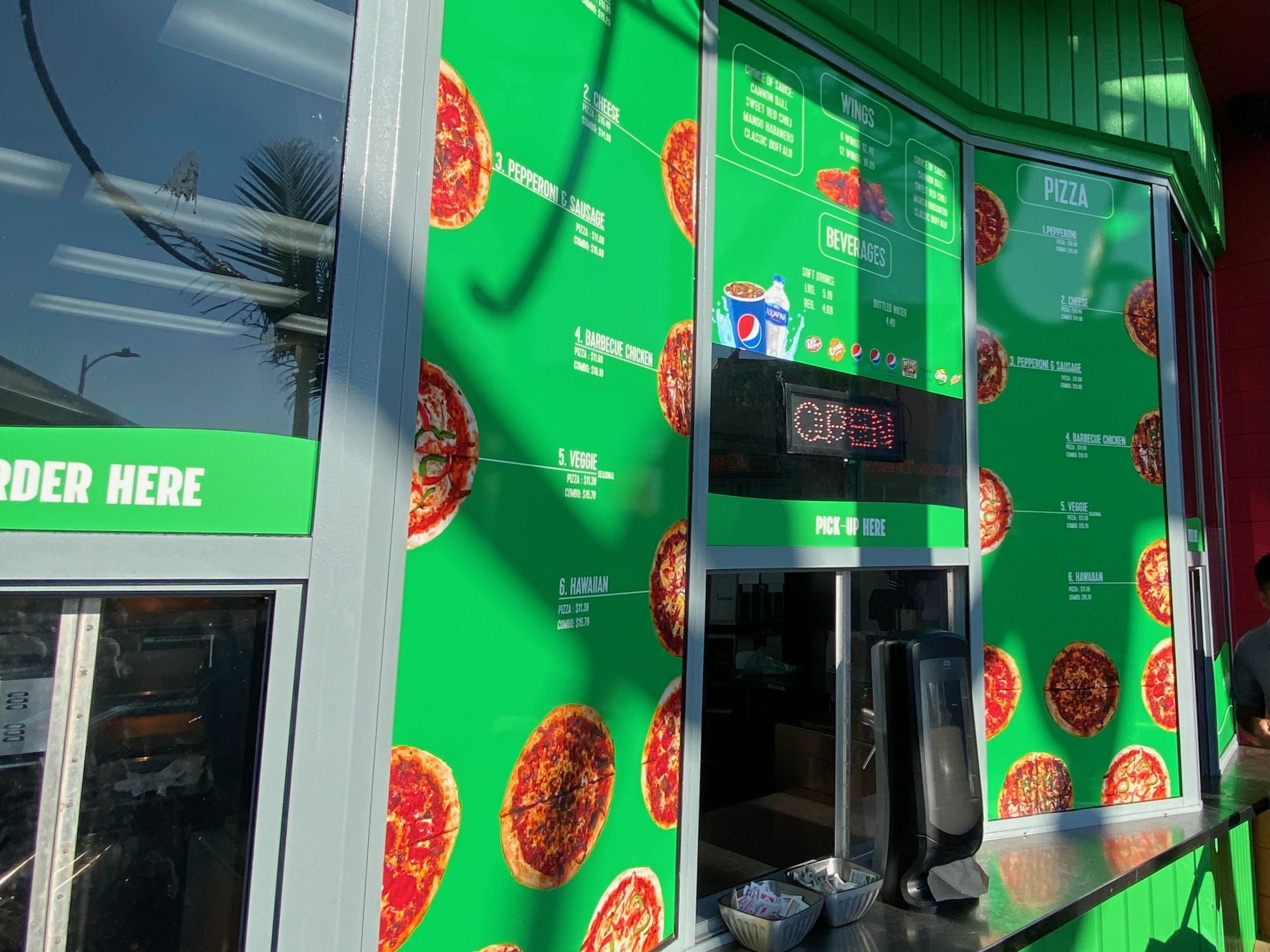Bright green menu wall of Mr. Nice Fry at Santa Monica Pier in Los Angeles, showing pizza options like pepperoni, cheese, veggie, Hawaiian and barbecue chicken, with the order window and pick-up window visible at the pier-side pizza stand.
