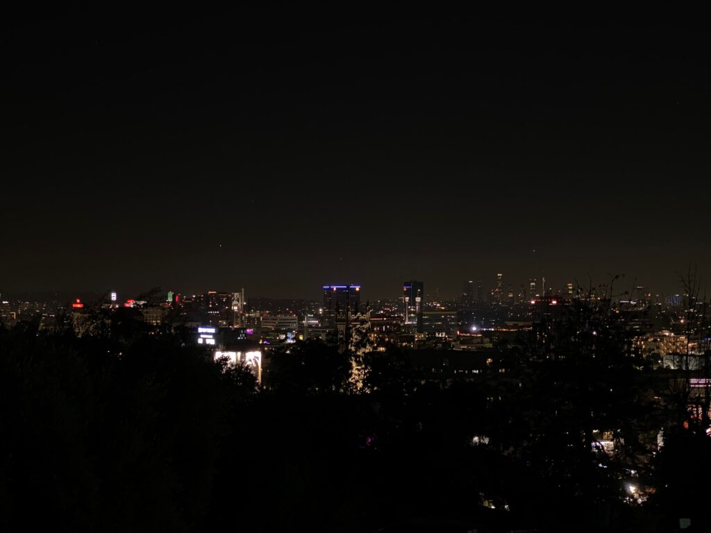 Night view from the Hollywood Hills overlooking illuminated Los Angeles, with city lights, downtown skyline, and dark hillside silhouettes in the foreground.