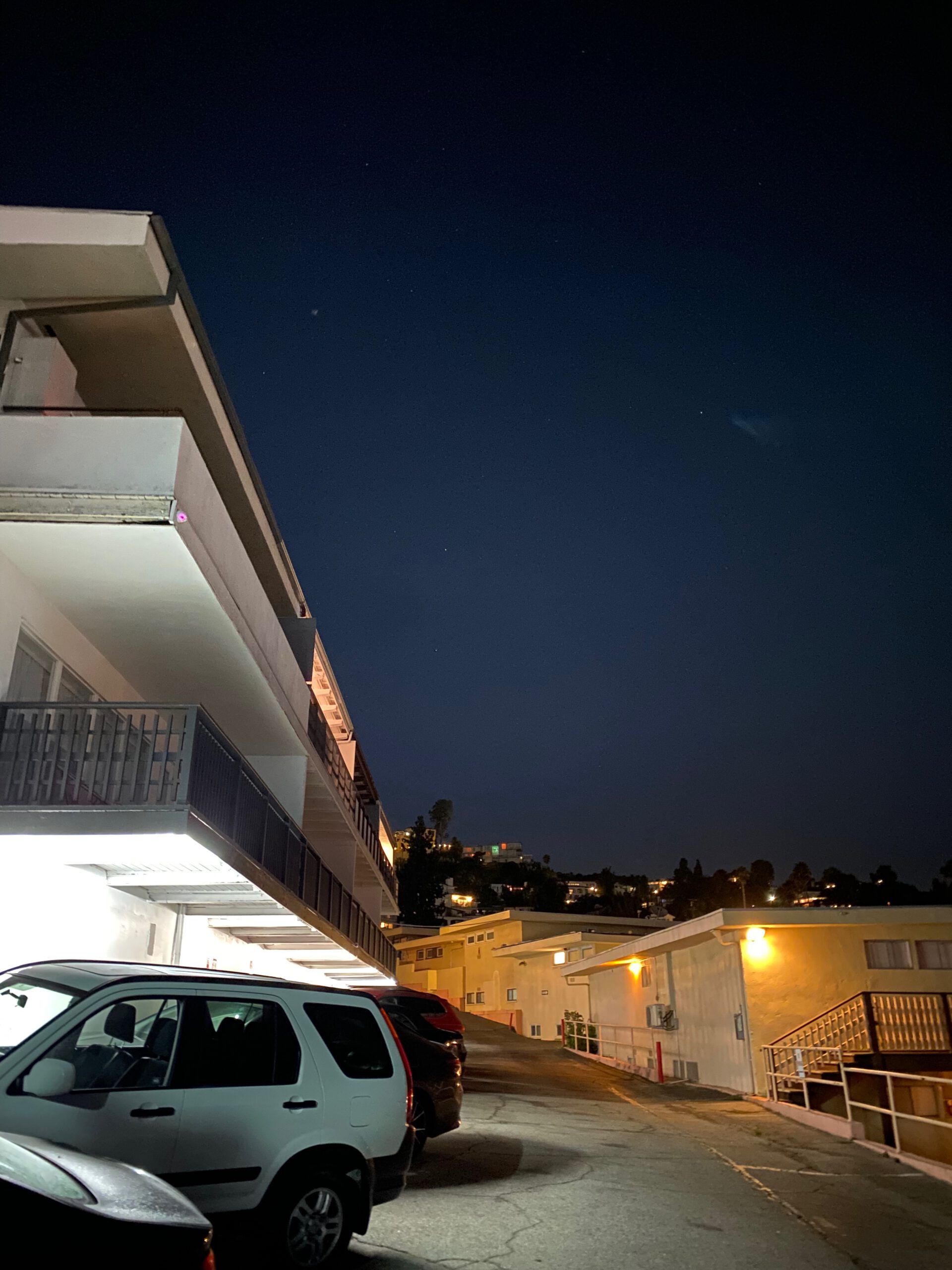 Night view of a Hollywood Hills hotel parking area with parked cars, illuminated buildings, and hillside homes visible in the distance under a dark sky.