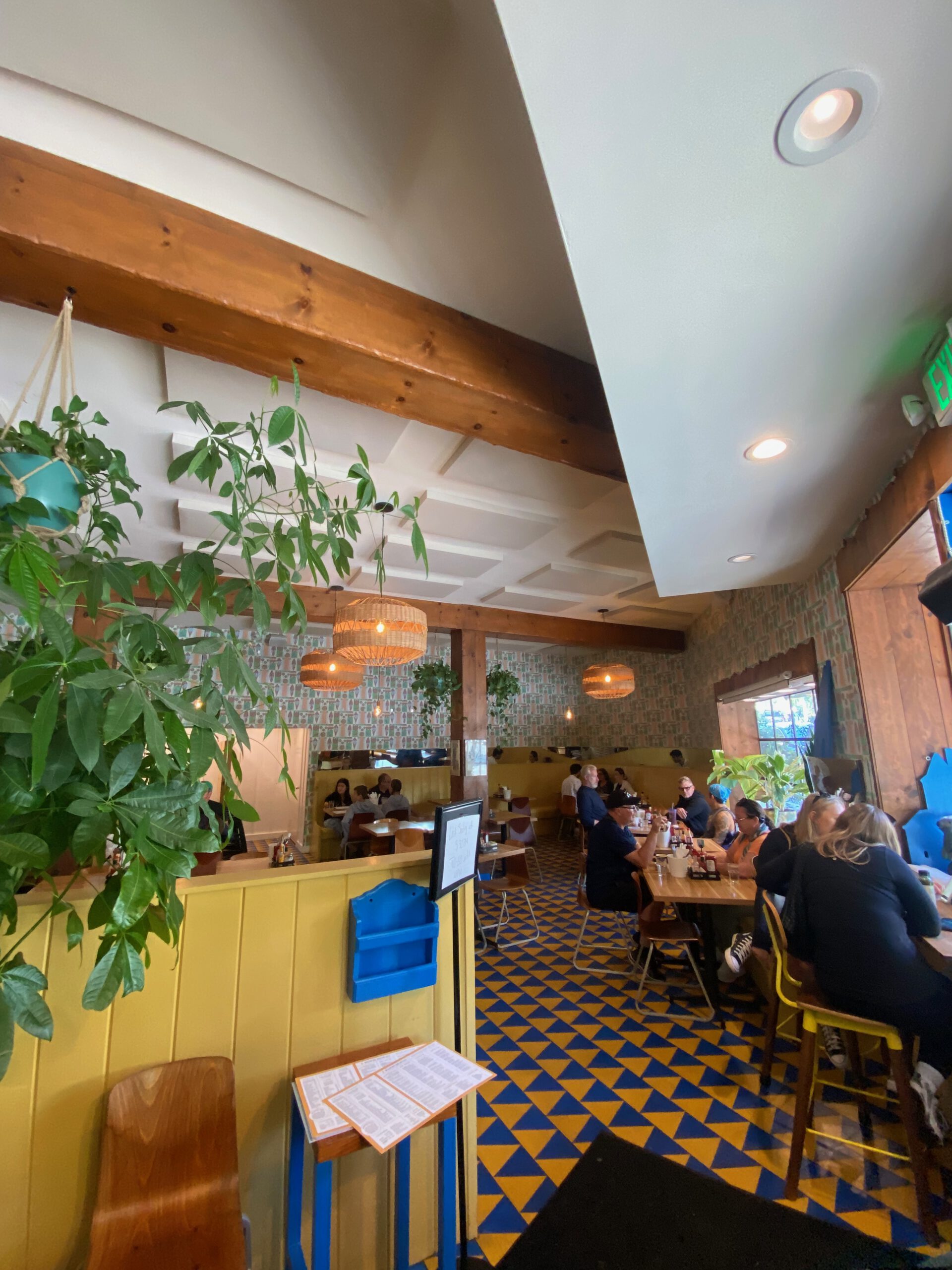 Interior of Beachwood Cafe Hollywood showing a cozy dining room with wooden beams, hanging plants, patterned blue-and-yellow tile floor, and guests enjoying breakfast.