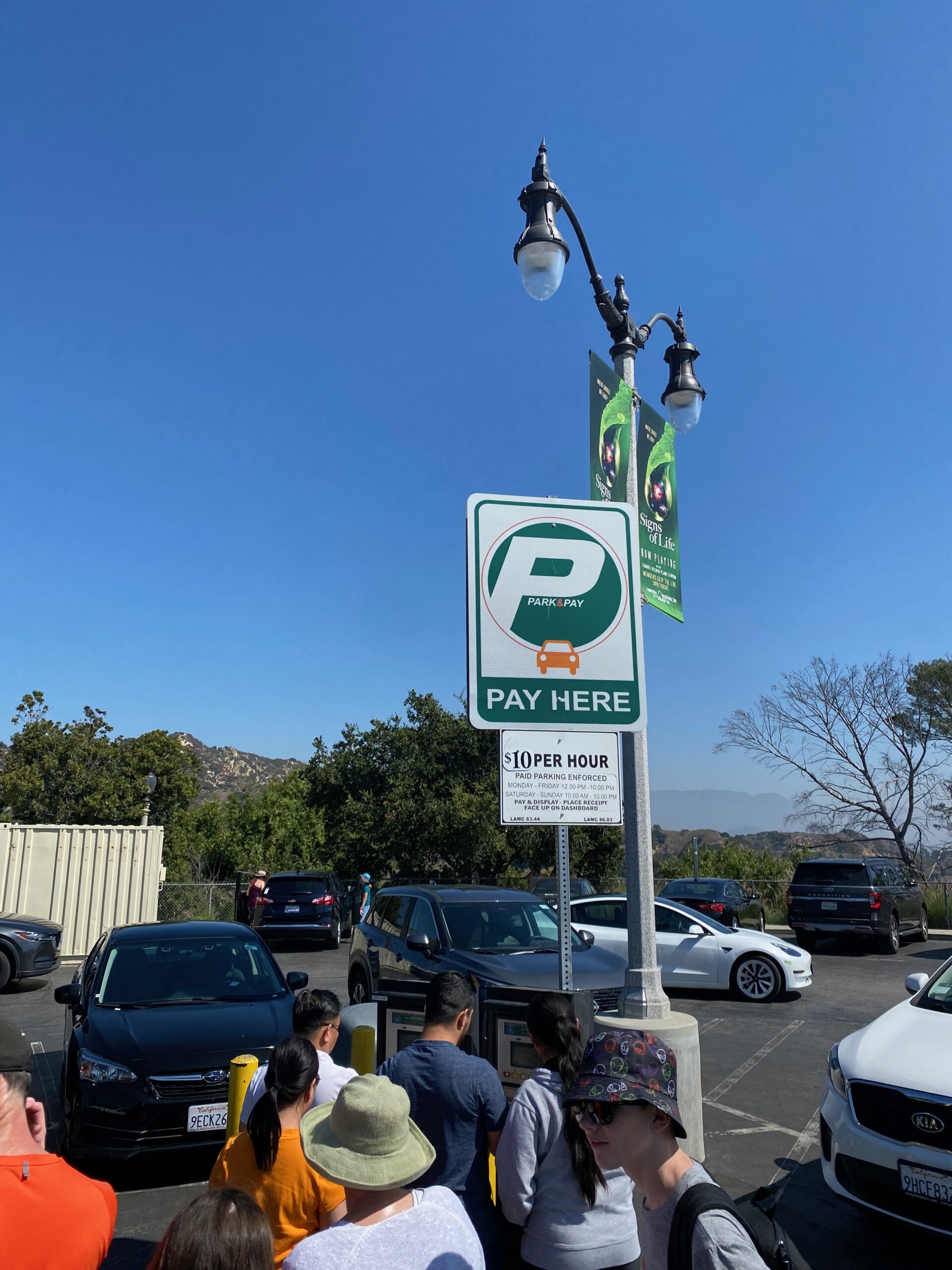 Visitors queuing at a Park & Pay parking meter directly in front of Griffith Observatory, with parked cars, paid parking signage showing $10 per hour, and clear blue skies overlooking the hills of Griffith Park in Los Angeles.
