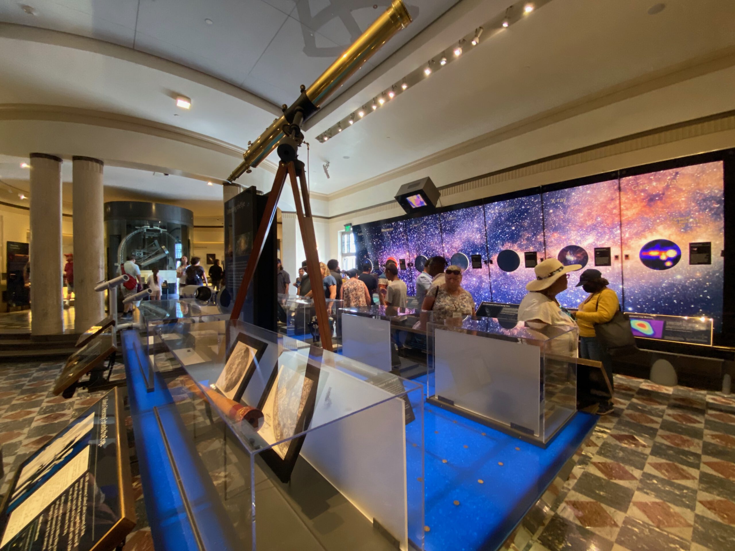 Historic telescope display inside Griffith Observatory with visitors viewing astronomy exhibits, glass cases, and a large cosmic timeline wall in Los Angeles.