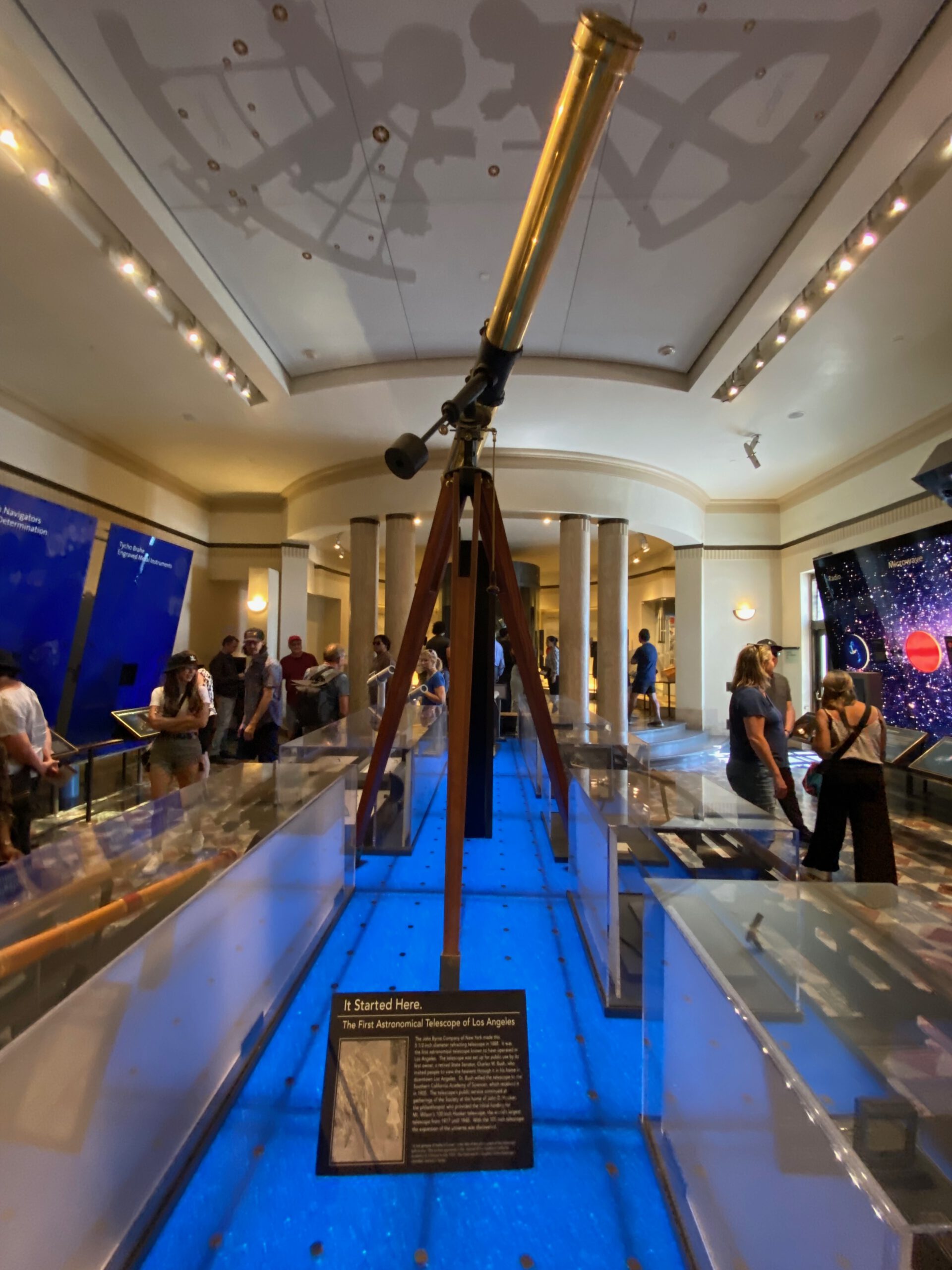 The first astronomical telescope of Los Angeles on display inside Griffith Observatory, shown on a wooden tripod with historic instruments and visitors exploring the gallery.